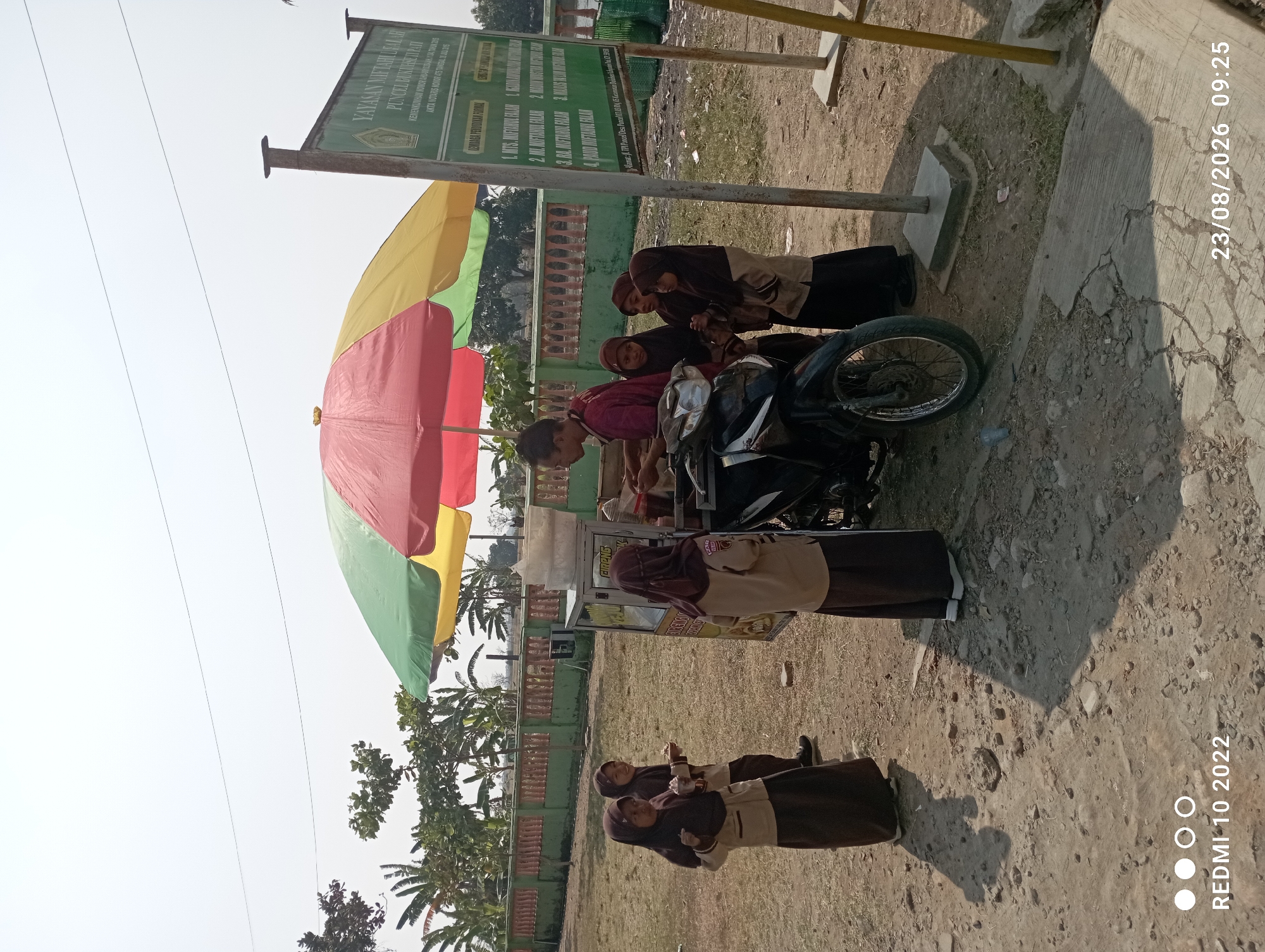 A street vendor is selling snacks from a cart attached to a motorcycle under a colorful umbrella. Several young girls in school uniforms are gathered around the cart, appearing to be customers. The scene takes place outdoors on a sunny day, with a fence and trees visible in the background. A sign for "YAYASAN MIFTAHUL FALAH" is prominently displayed to the right, indicating the location is likely near an educational institution in Dukuhseti, Indonesia. The ground is a mixture of dirt and cracked pavement.