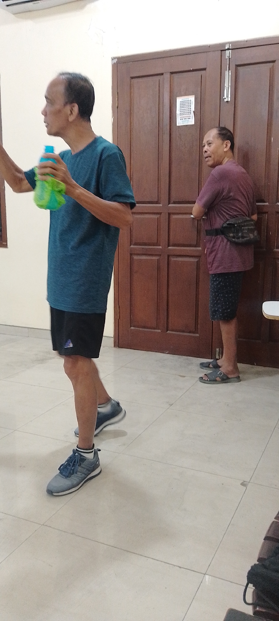 In Kudus, Indonesia, two adult males are standing on a tiled floor. The man on the left is wearing a blue t-shirt, black shorts, and grey athletic shoes. He is holding a green and blue water bottle in his right hand. The man on the right is wearing a maroon t-shirt, patterned shorts, and dark sandals. He has a dark fanny pack around his waist. They are standing in front of a double wooden door with a vertical metal latch. The wall behind the door has a small, square white paper attached to it. The room appears to be indoors, with a tiled floor and a light-colored wall.