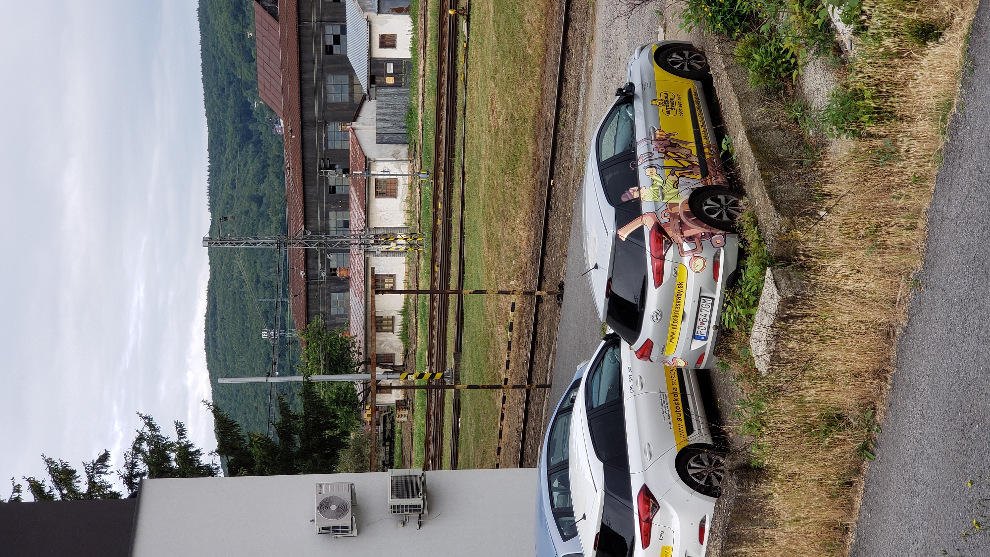 In Prešov, Slovakia, two white Hyundai i20 cars, marked with "AUTOSKOLA SVABY.SK" and cartoonish graphics, are parked next to a railway line. The car on the right bears the license plate "PO647GM." In the background, a series of low-rise industrial buildings with rusty roofs and windows are visible, beyond which a dense, tree-covered hill rises under an overcast sky. Two air conditioning units are mounted on the white exterior wall of a building in the foreground on the left. The scene is framed by dry grass and concrete barriers.