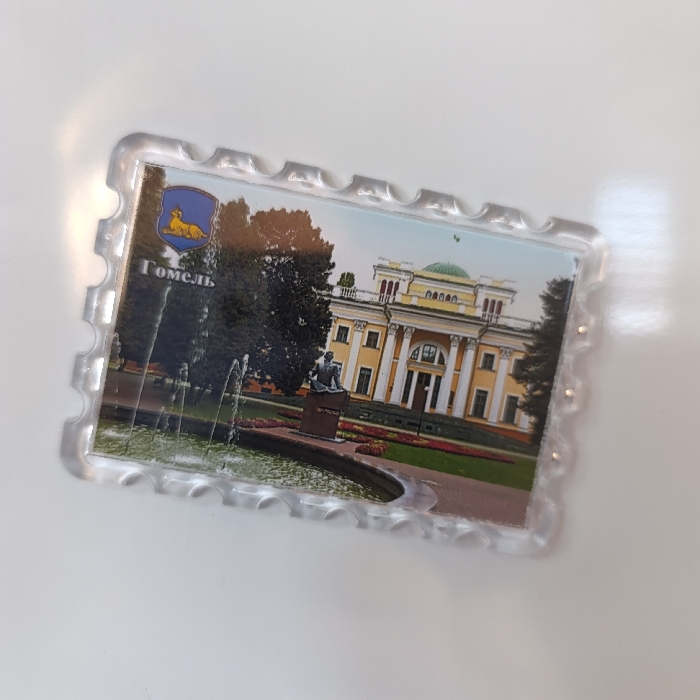 A clear plastic souvenir magnet, rectangular with a serrated border resembling a postage stamp, is depicted lying on a white, reflective surface. The magnet features a color photograph of a prominent, multi-story palace characterized by a yellow facade, white classical columns supporting a central portico, and a distinct green domed roof. In the foreground, a circular fountain actively sprays water, positioned near a dark, seated figure statue atop a rectangular pedestal. The immediate surroundings consist of manicured green lawns interspersed with beds of red and orange flowers. Dense green trees frame the palace on both sides. In the upper left quadrant of the photograph, a blue heraldic shield with a golden reclining lion is visible, beneath which the white Cyrillic text "Гомель" (Gomel) is printed. Reflections of ambient light are visible on the magnet's plastic surface and the underlying white background. The scene depicted on the magnet is of the Rumyantsev-Paskevich Palace in Gomel, Belarus.