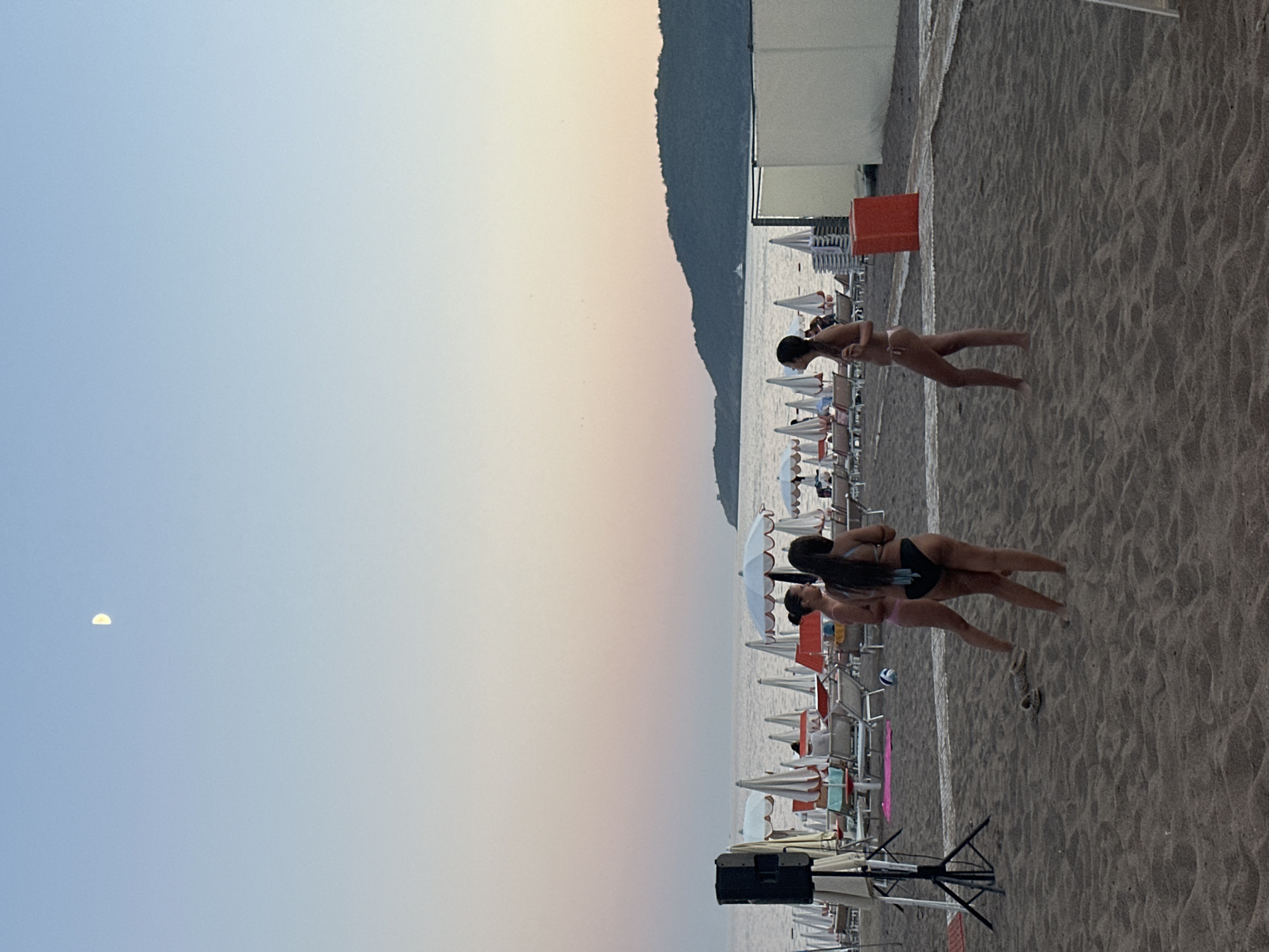 A group of three young women, dressed in bikinis, are standing on a sandy beach in Boreti, Montenegro. Two of the women are facing away from the camera, while one is partially turned to the side. They appear to be engaged in a casual conversation or activity.
In the background, rows of beach chairs and umbrellas are set up, suggesting a recreational area. Beyond the beach, the sea stretches out to the horizon, with a gentle slope of land visible in the distance. The sky is a soft gradient of blue and pink, indicating either dawn or dusk. A crescent moon is visible in the upper left quadrant of the sky. A portable speaker on a tripod is positioned on the left side of the frame, possibly for music. A red bin is visible on the right side of the frame.