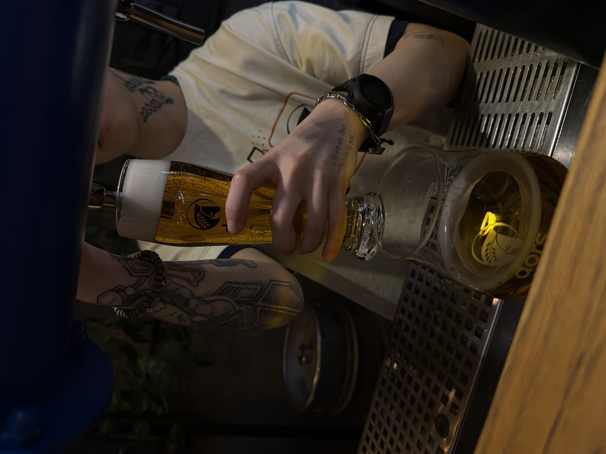 A person is pouring beer from a tap into a glass in Yerevan, Armenia. The person has tattoos on their arm and is wearing a watch. The glass is being filled with a light golden beer that forms a white foamy head. The beer is being poured on top of a metal drip tray, positioned to catch overflow. The area has wooden paneling.