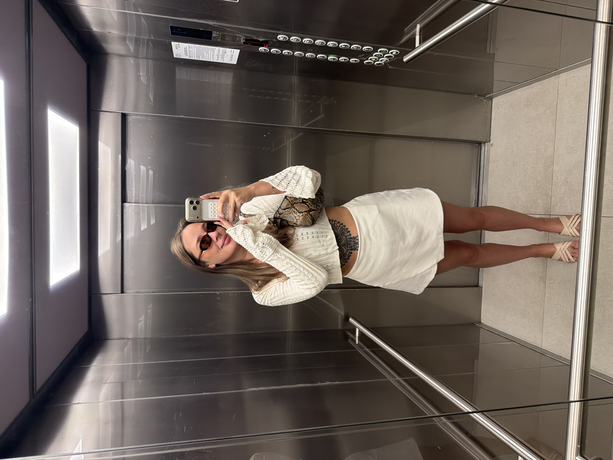 A woman is standing in an elevator, taking a selfie. She is wearing sunglasses, a white cropped cardigan with intricate detailing, and a white wrap skirt. She has a snake-print purse slung over her shoulder and a large tattoo on her stomach. She is wearing light-colored heeled sandals. The elevator is modern and metallic, with a digital panel of buttons on the right wall. The floor is tiled. The lighting appears to be from overhead fluorescent lights. The image does not contain any explicit information to indicate the city of Moscow, Russia.