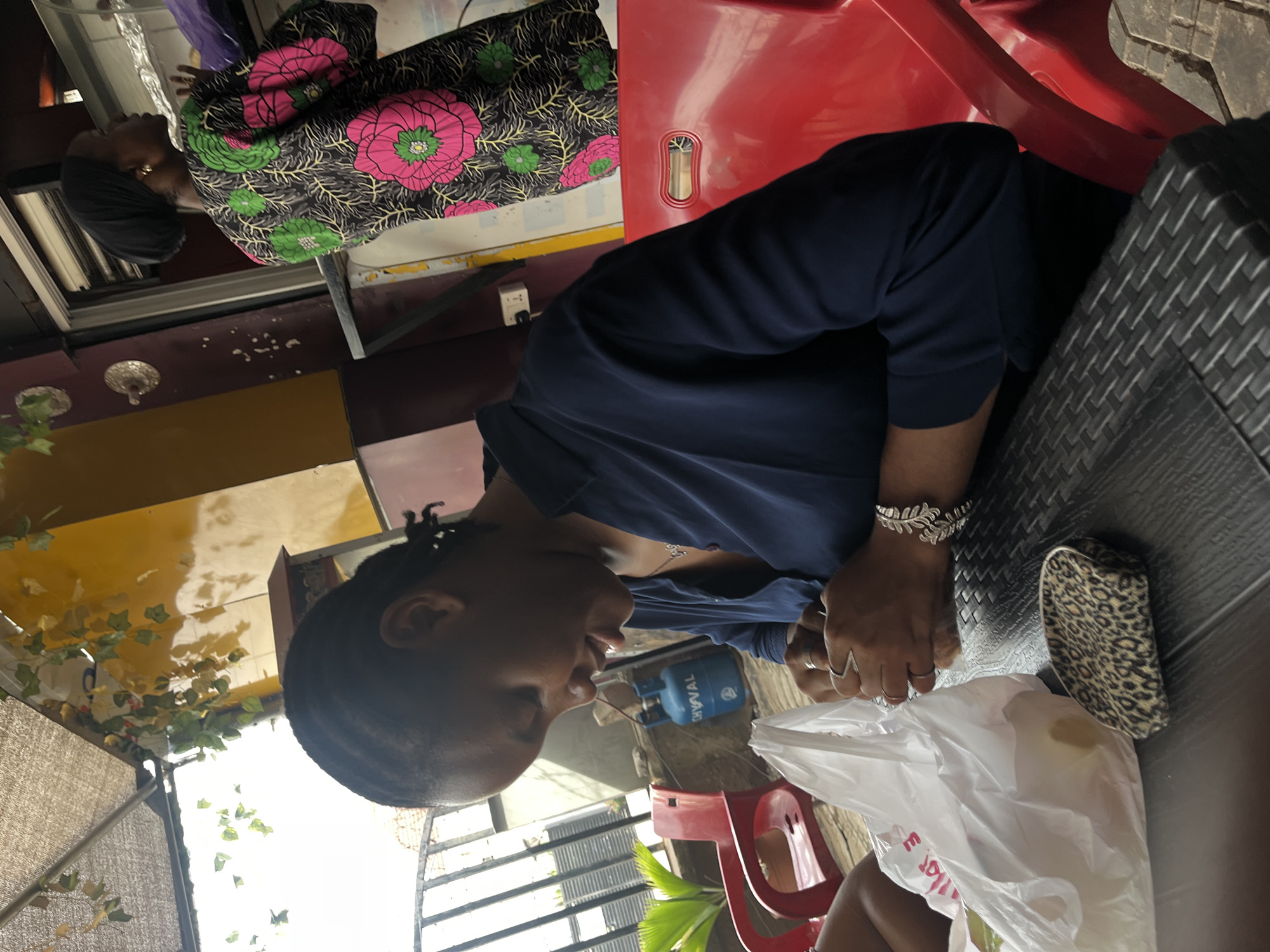 The image shows two Black women sitting at a table outdoors in Karum, Nigeria. One woman is seen from the waist up, wearing a brightly patterned Ankara dress. The second woman, closer to the camera, is wearing a navy blue shirt and a silver bracelet. She is seated at a black wicker table next to a red chair. A white plastic bag and a leopard print purse are also on the table. Behind her, a blue gas cylinder and a red chair are visible, along with a glimpse of outdoor scenery with plants and a fence. The overall setting suggests an informal outdoor dining area.