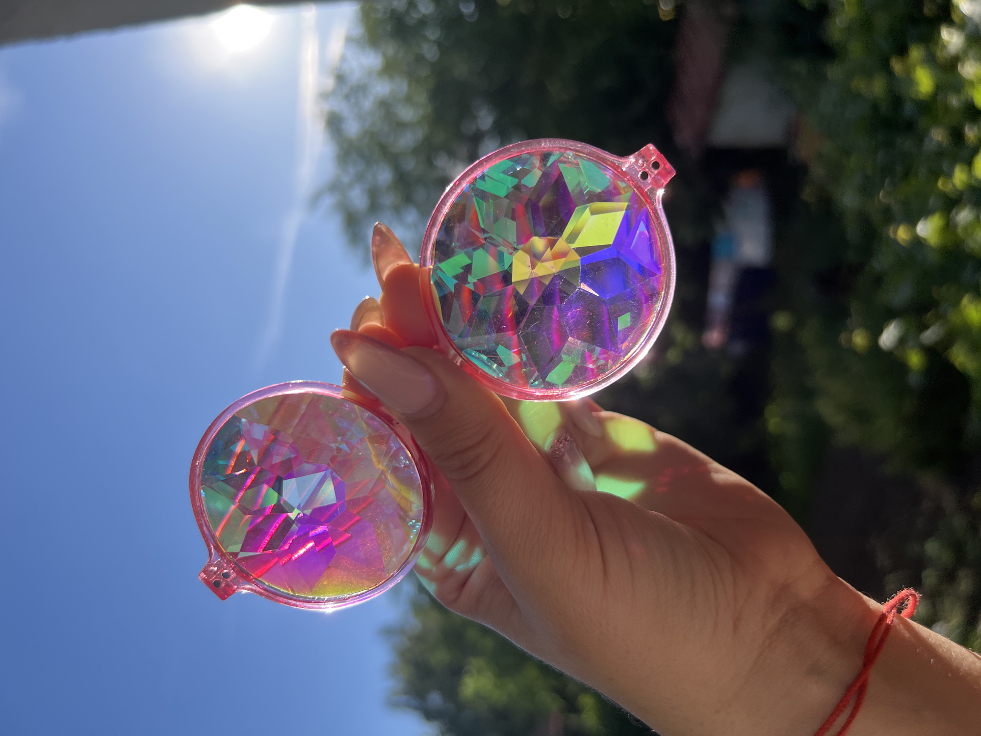 A hand holds a pair of pink kaleidoscope glasses with multi-colored geometric lenses up to a clear blue sky. The sun is bright and shining, creating lens flare on the right side of the image. In the background, out-of-focus green trees and hints of buildings suggest an outdoor setting, possibly in a residential area. A thin red string is tied around the wrist of the person holding the glasses. The image appears to have been taken during the day.