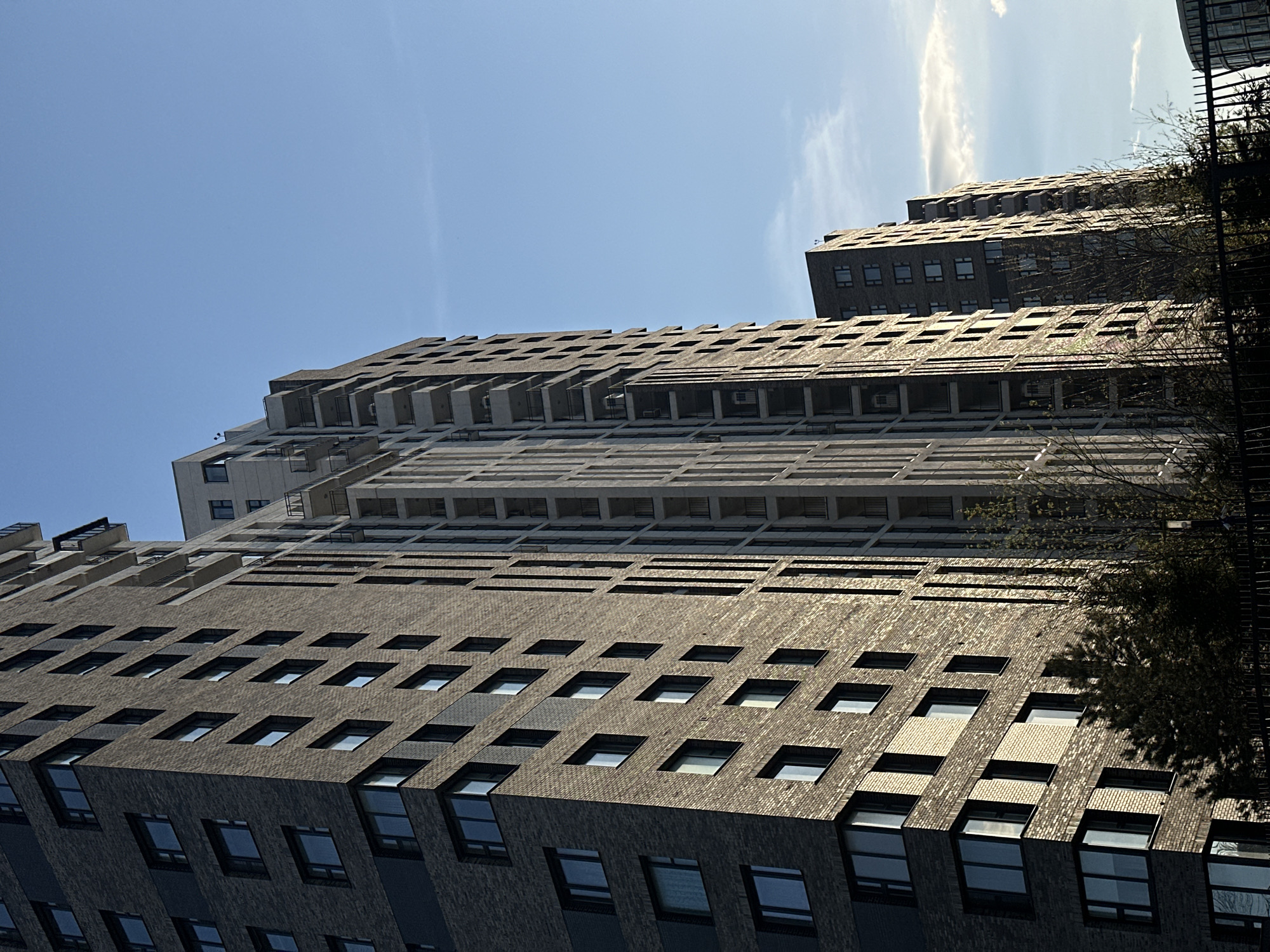 The image shows a tall building in Moscow, Russia, viewed from a low angle. The building has a gray brick facade with evenly spaced square windows. The top of the building has a series of balconies or overhangs. The sky is blue with a few wispy clouds. Part of another building is visible in the background, partially obscured by trees.