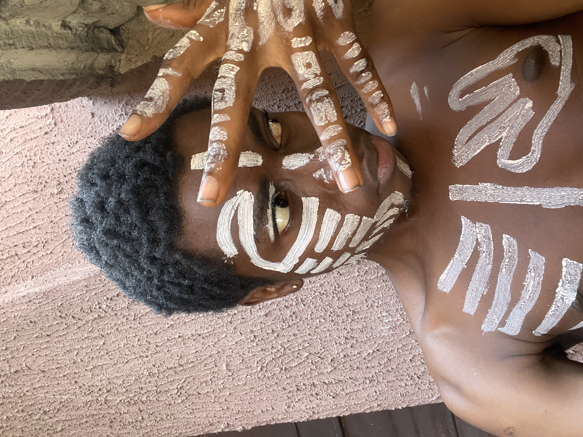 An individual with dark skin and dark, tightly curled hair is depicted lying down, with their head positioned towards the left of the frame and their body extending to the right. Their eyes are open and directed upwards. The person's face, hands, and upper torso are adorned with intricate white body paint, suggesting a cultural or ceremonial event taking place in Onitsha, Nigeria.
On the face, bold white lines frame the eyes, with strokes extending from the inner corners and forming distinct rectangular and linear patterns on the cheeks and forehead. A thick white line runs across the bridge of the nose. The individual's hands, with natural, unpainted fingernails, are positioned near their face, also displaying various white linear and block patterns on the back of the fingers and knuckles. On the upper chest and shoulder area, thick, sweeping white strokes form prominent designs, accompanied by several thinner vertical lines further to the left.
The setting appears to be indoors or in a shaded outdoor area, with a light brown, highly textured surface visible as the background, underneath and behind the individual. The lighting is soft, indicating neither harsh sunlight nor deep shadows. The overall mood conveyed is one of contemplation or solemnity, consistent with a traditional ritual or artistic expression. No visible text is present in the image.