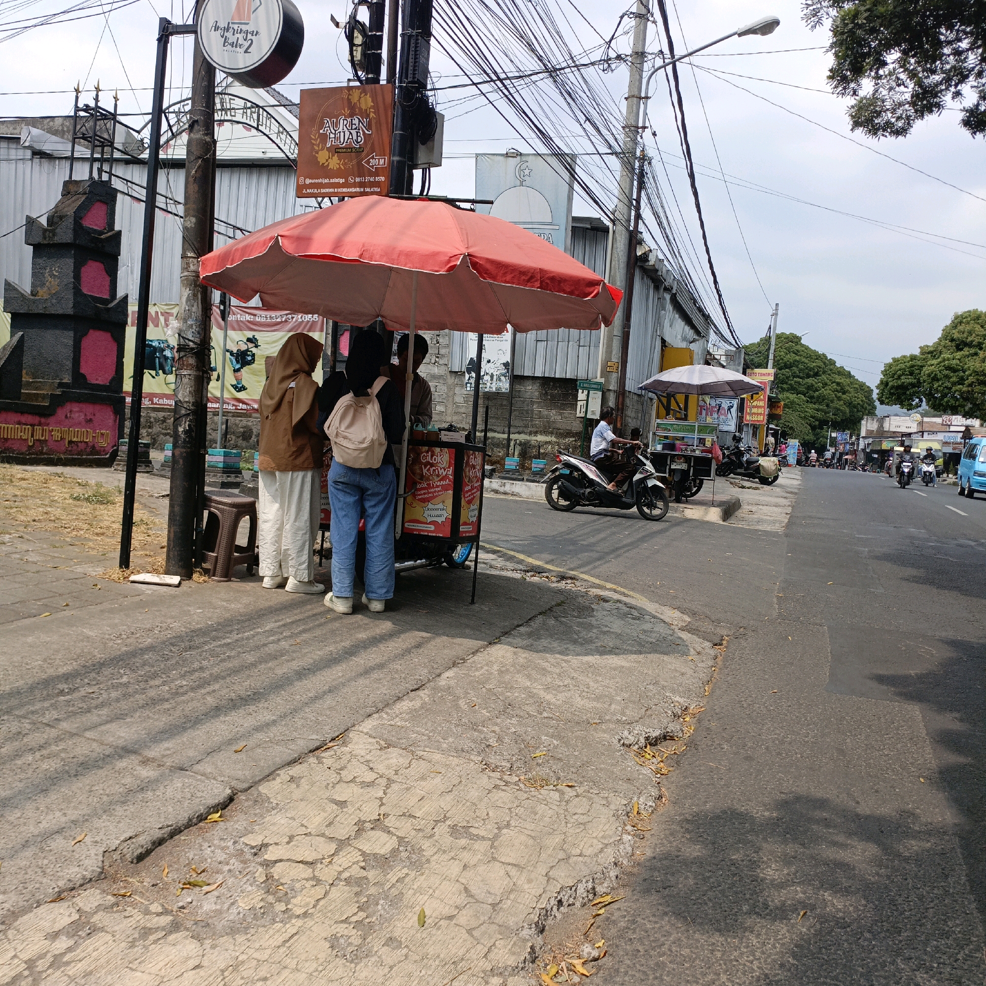 This is a daytime, medium-angle, eye-level shot of a street scene in Salatiga, Indonesia, featuring a food stall and surrounding urban environment. The foreground is dominated by a red umbrella covering a food cart. Three people are visible near the cart. Two individuals, likely women, are standing, and one is wearing a brown hijab and white pants, while the other is wearing blue jeans and a backpack. A third person, likely a vendor, is standing behind the cart, facing them. The cart itself has colorful signage, advertising food items.
To the left of the food stall, a dark, ornate structure with pink accents stands next to a utility pole adorned with multiple signs. The signs include one for "Angkringan Babe 2" and another for "AUREN HIJAB." Further to the left, a sidewalk is visible, with scattered dry leaves.
The middle ground shows a street with various motorbikes. One motorbike is parked, while another is being ridden by a person in a blue shirt. A second, smaller food stall with a white umbrella is visible further down the street. The background consists of buildings, trees, and a cloudy sky. The road surface is asphalt, with a visible yellow line marking the lane. The overall scene depicts a typical street-level commercial activity in an Indonesian city.