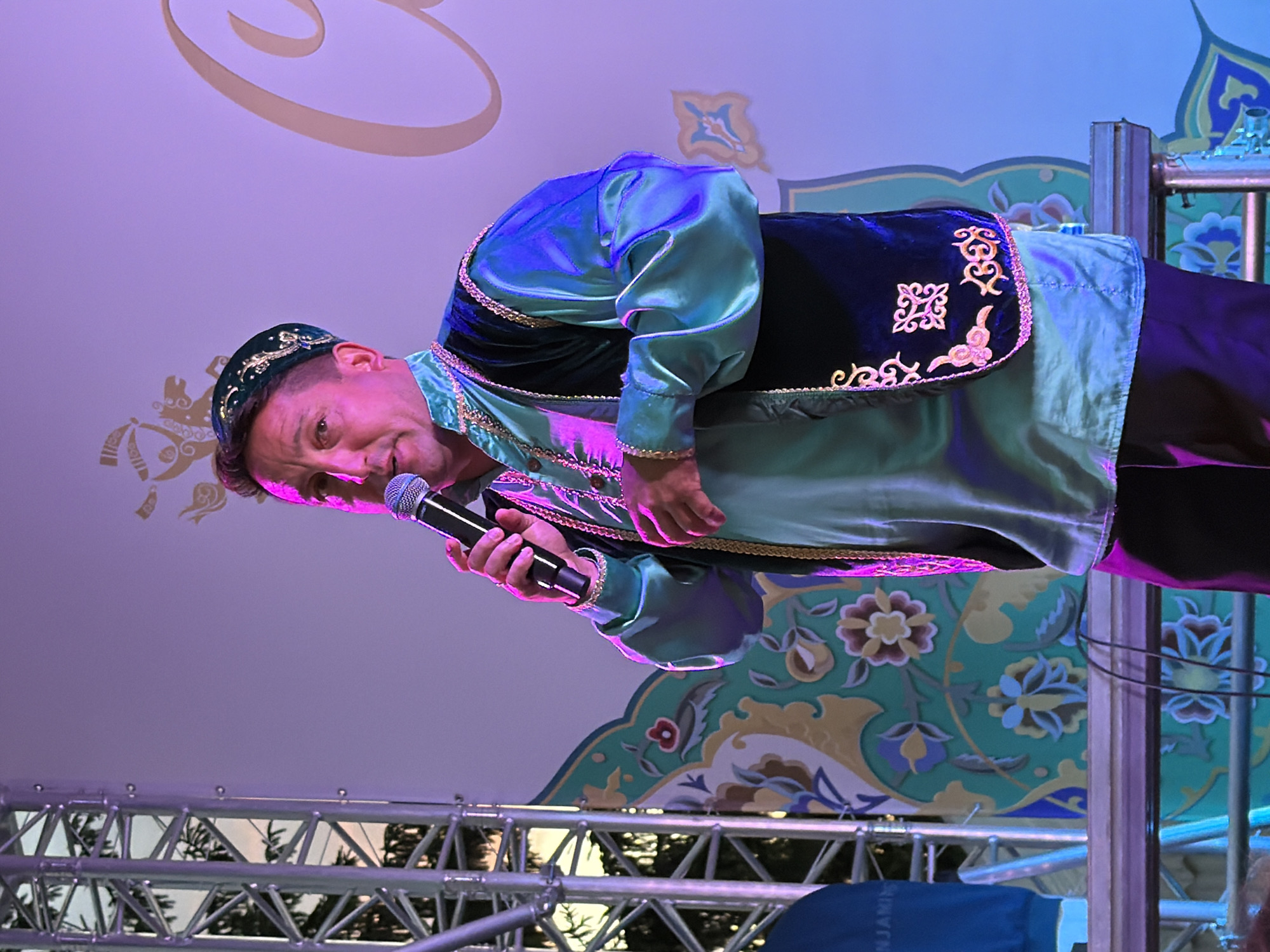 A man in traditional attire stands on a stage, holding a microphone and speaking. He wears a green cap adorned with gold embroidery, a shimmering aqua-colored shirt with puffed sleeves, and a dark vest with gold trim and intricate patterns. The man is lit by purple and blue stage lights, which cast colored hues on his clothing and skin. Behind him, a large backdrop features a light-colored design with floral motifs in teal and gold, and a large, ornate cursive letter. To the left of the man, a metal truss structure is visible, partially obscured by the stage lights. The man appears to be addressing an audience, though they are not visible in the frame.