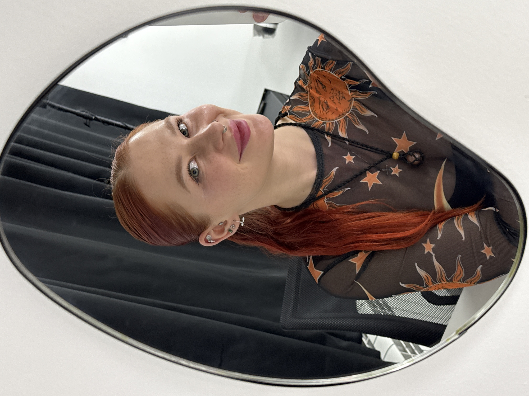 A woman with red hair is reflected in an oval mirror. She is wearing a black mesh top with orange sun and star patterns. The mirror is hanging on a white wall. Behind her, in the mirror's reflection, is a black curtain and a black mesh chair. There is no information to suggest the image was taken in Rostov-on-Don, Russia.