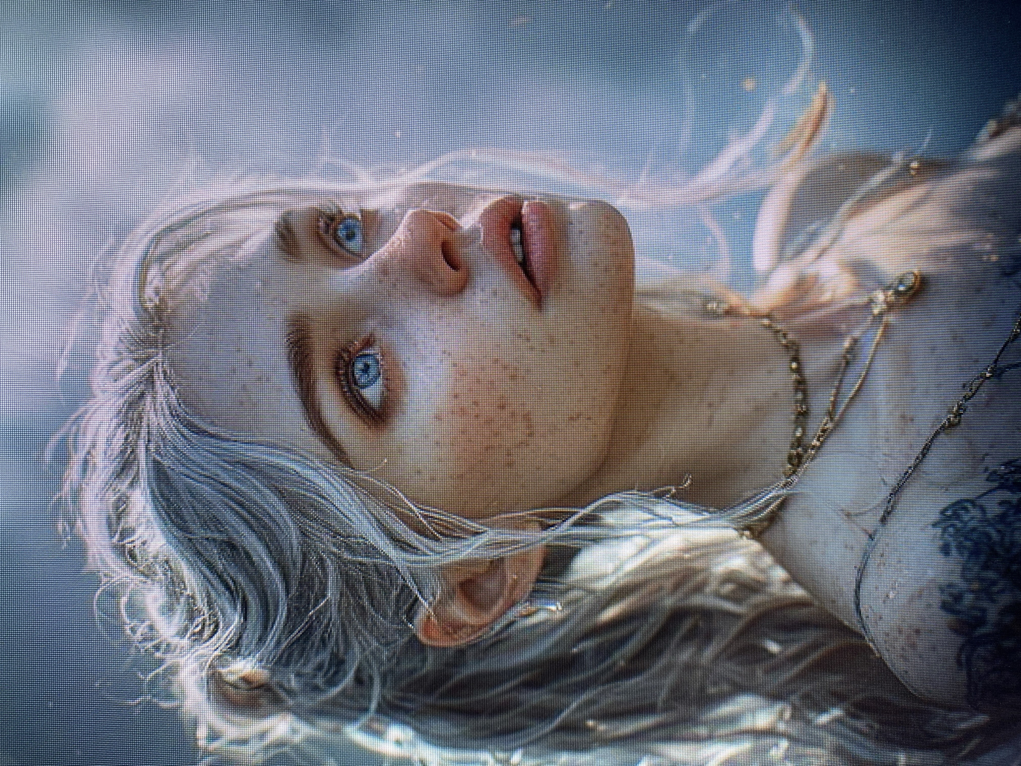 This media file is a close-up, overhead shot of a young person submerged in water. The subject has fair skin with freckles, blue eyes, and light-colored hair that is spread out around their head. They are looking upwards, with their mouth slightly open. The subject is wearing a gold necklace with intricate detailing. A portion of their torso is visible, revealing a dark tattoo on their right shoulder. The background is a soft, blurred blue, suggesting an aquatic environment, possibly a pool or a natural body of water. The lighting is diffused, creating a dreamlike or ethereal quality to the image. The overall mood appears serene or contemplative. No text is visible in the image.