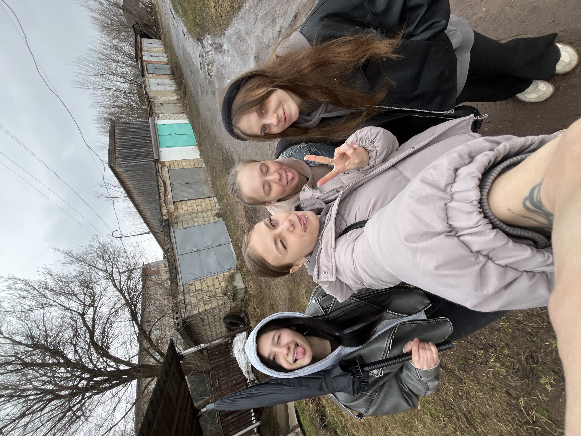 Four young women are posing for a selfie in an urban or semi-urban environment. The selfie taker's arm is visible on the right side of the frame, bearing a tattoo. The top of the head of one of the women is wearing a black headband. The woman in the middle is wearing a light purple/gray puffer jacket and is extending her hand with two fingers up in a peace sign. The lower woman is wearing a black leather jacket with a gray hood, and is holding an umbrella. Behind the women, a row of garages with gray and teal doors are visible. A bare tree stands to the left of the garages. The ground appears damp. The image was likely taken in Фоки, Russia.