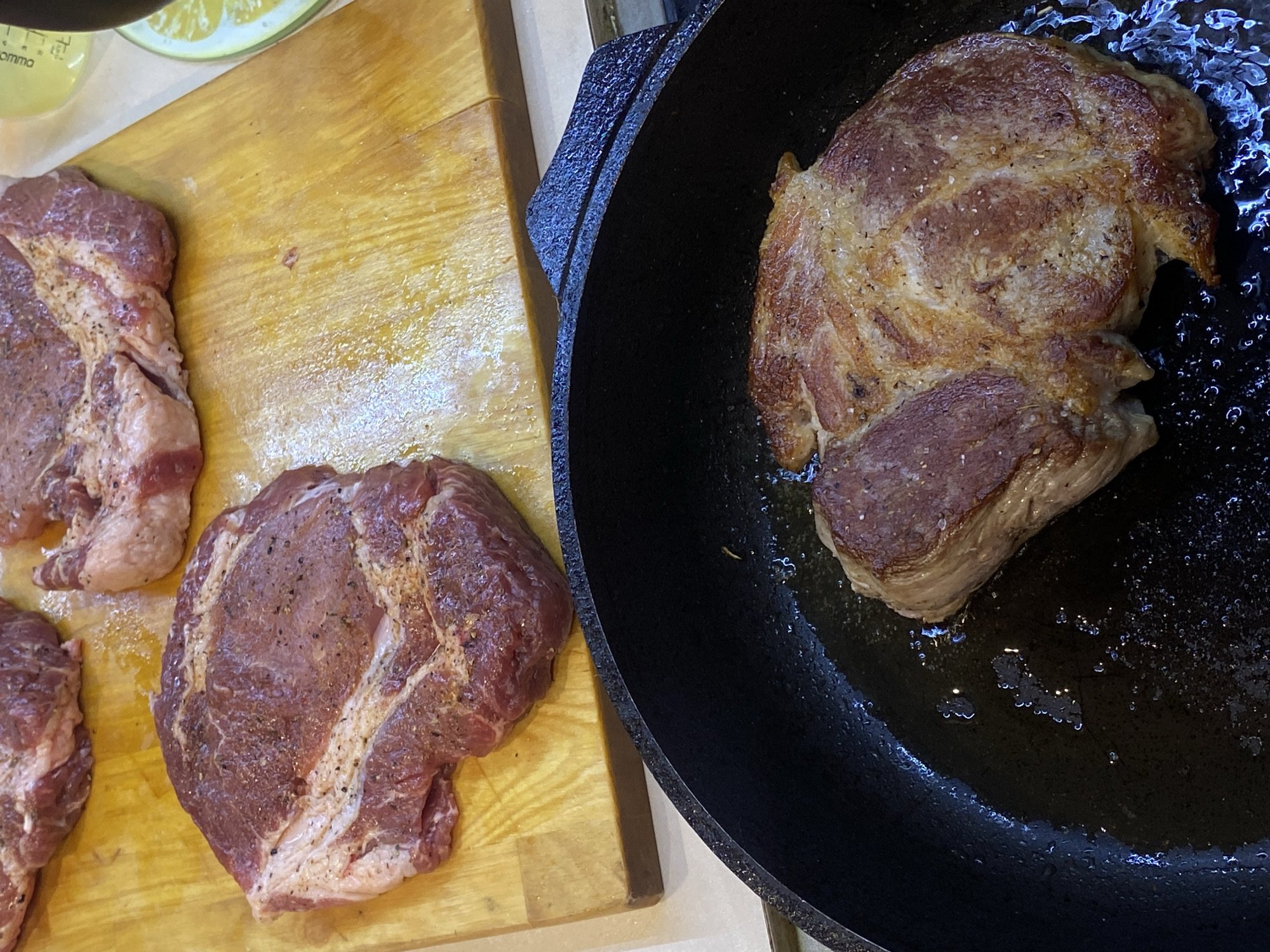 The image shows raw steaks on a wooden cutting board alongside a single steak cooking in a black cast iron skillet. The steaks are red with visible marbling and seasonings, suggesting they might be pork or beef. The cooked steak has a golden-brown crust, indicating it is being seared. There are no people visible in the image. The location is not identifiable as Votkinsk, Russia, or any specific location.