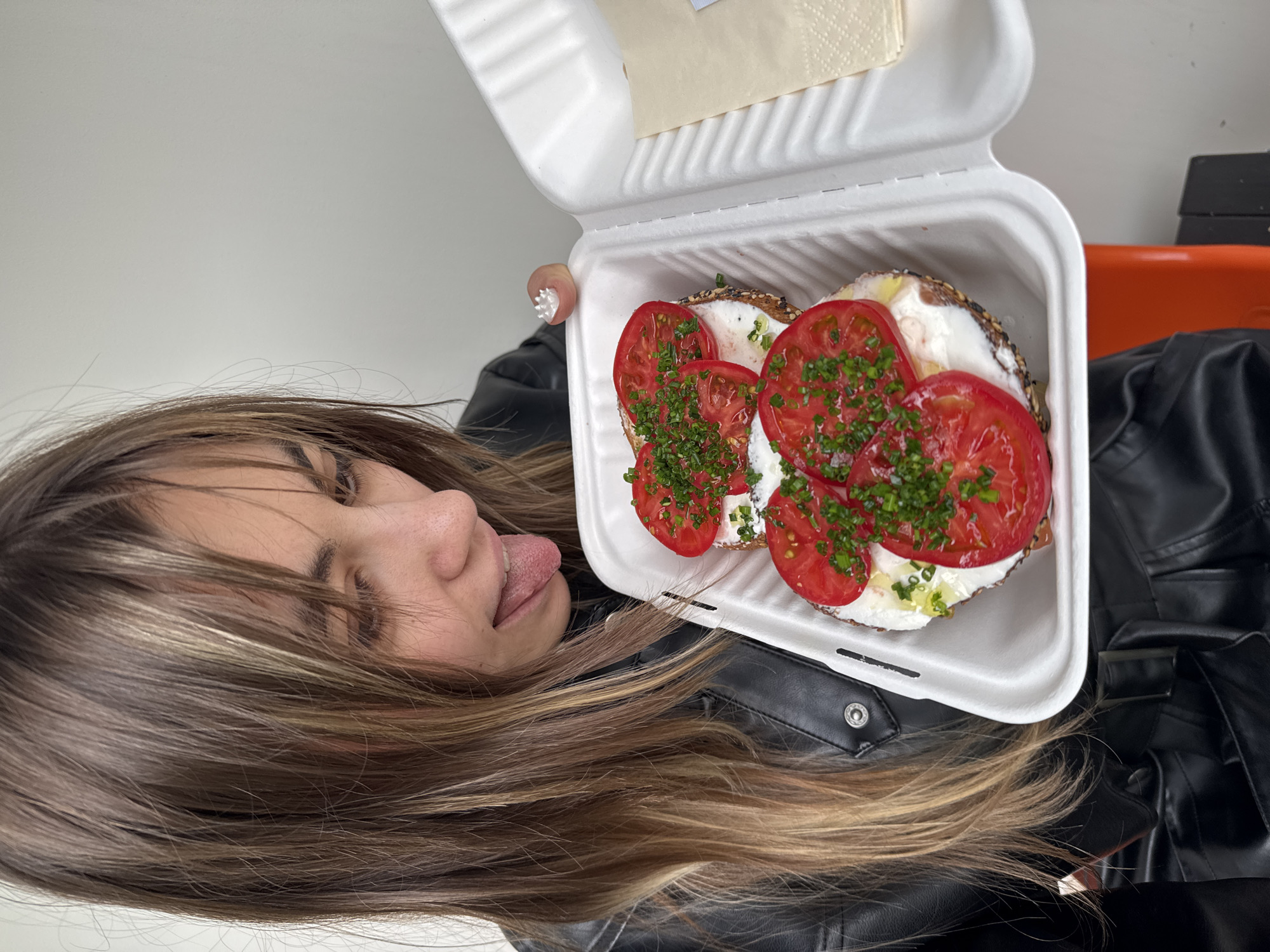 A young woman with long, brown and blonde-streaked hair, wearing a black leather jacket, is looking down at a white takeout container filled with two open-faced bagels. The bagels are topped with cream cheese, sliced tomatoes, and chopped green onions. Her tongue is sticking out as she looks at the food. In the background, a white wall is visible. The scene is outdoors, possibly on a street in Moscow, Russia.