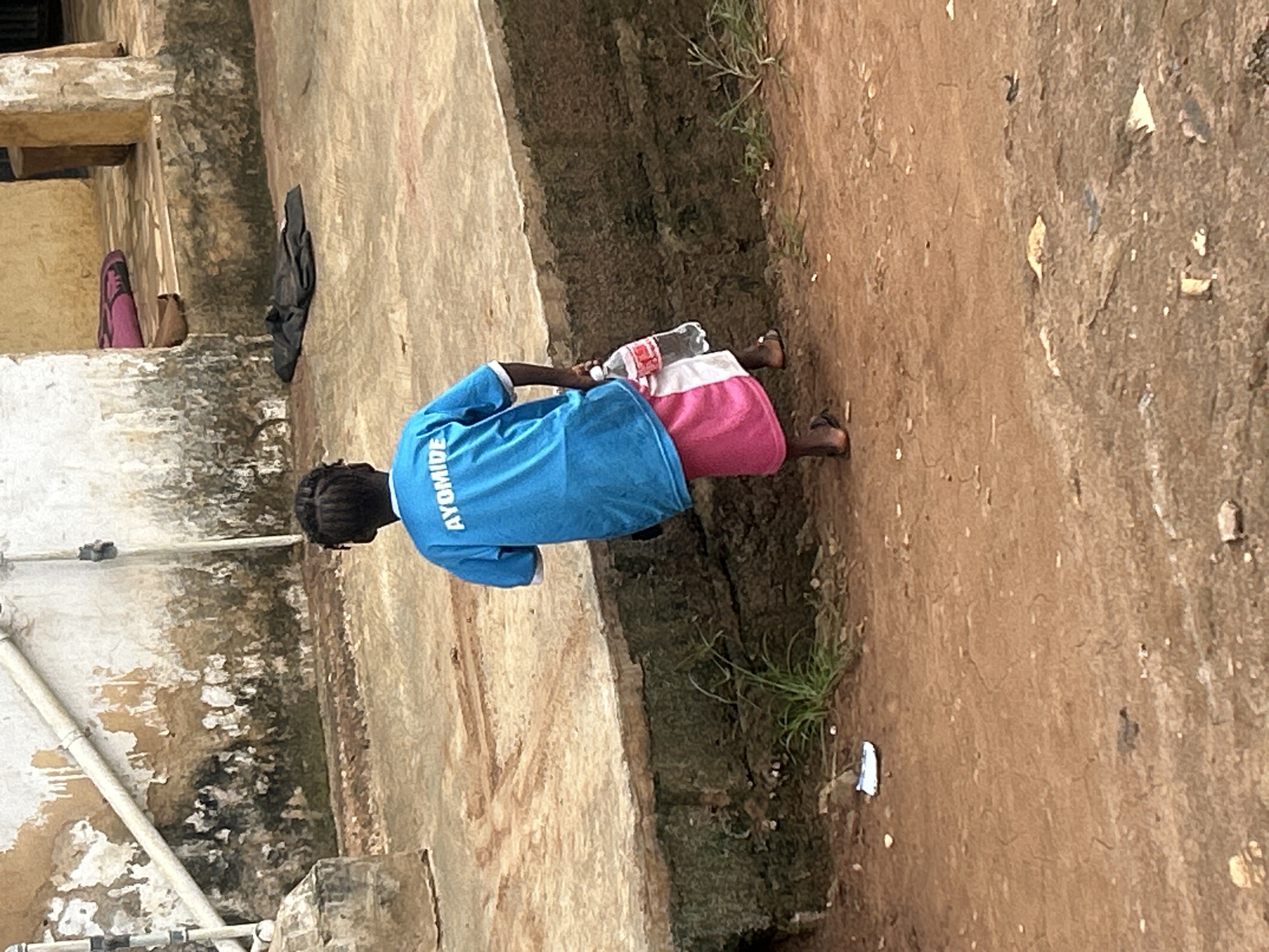 A young child, seen from behind, walks on a dirt path in what appears to be an outdoor, possibly impoverished, setting. The child wears a bright blue t-shirt with white lettering that reads "AYOMIDE" and a pink and white skirt. They are carrying a clear plastic bottle of water in their right hand. The child's hair is dark and styled in braids. The ground is a mix of reddish-brown dirt, with some scattered debris and patches of sparse green vegetation. In the background, a rough concrete wall with peeling paint and some visible pipes suggests a rustic or dilapidated structure. A dark, folded garment lies on the ground near the wall. The overall impression is a scene from a rural or informal urban area in Ikorodu, Nigeria.