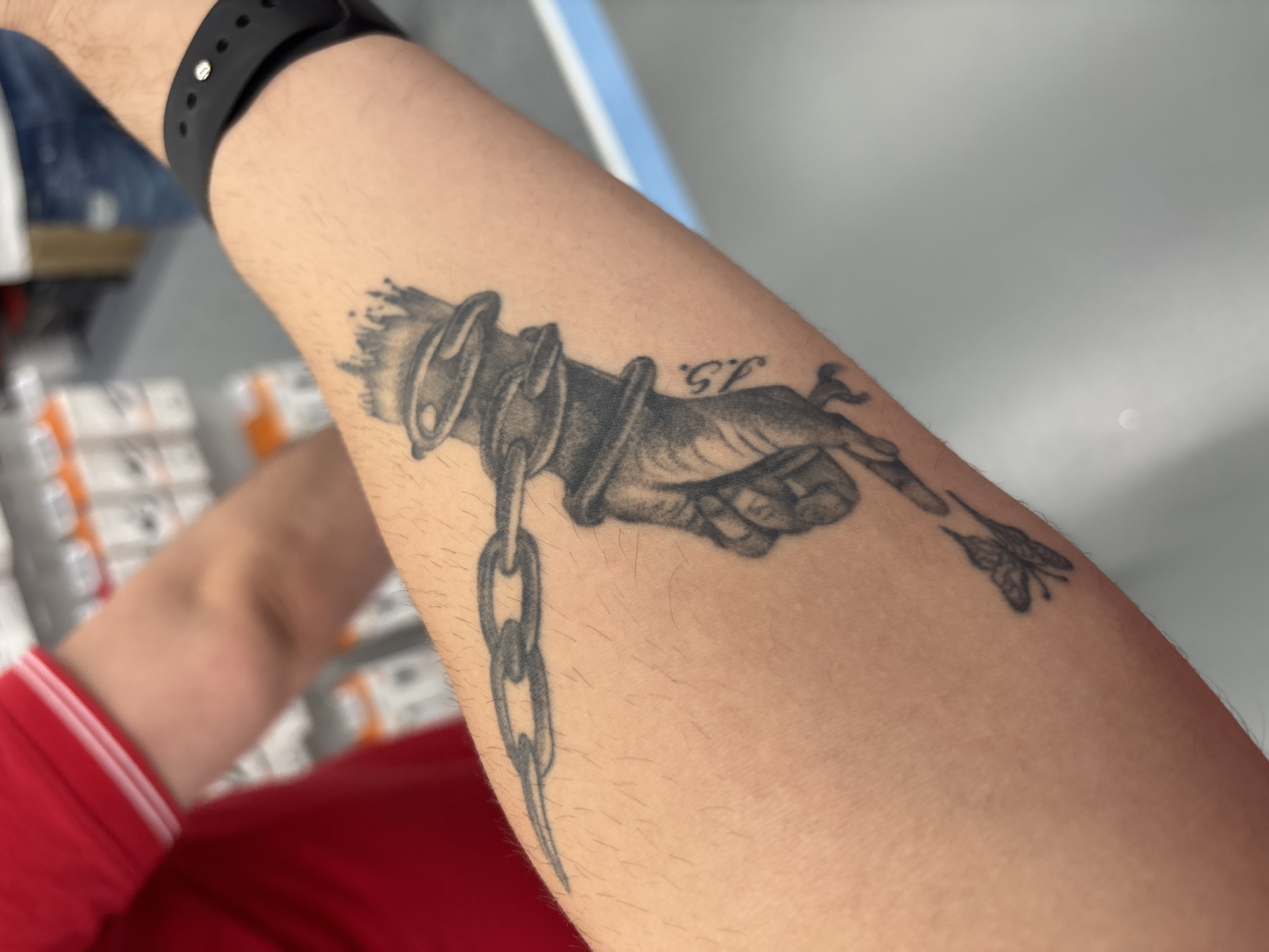 The image shows a forearm with a tattoo of a hand chained and holding a flower. The forearm has a black smart watch on the wrist. The person is wearing a red shirt. The background is blurry and shows items on shelves. No information is provided regarding the location in the city of Lovech, Bulgaria.