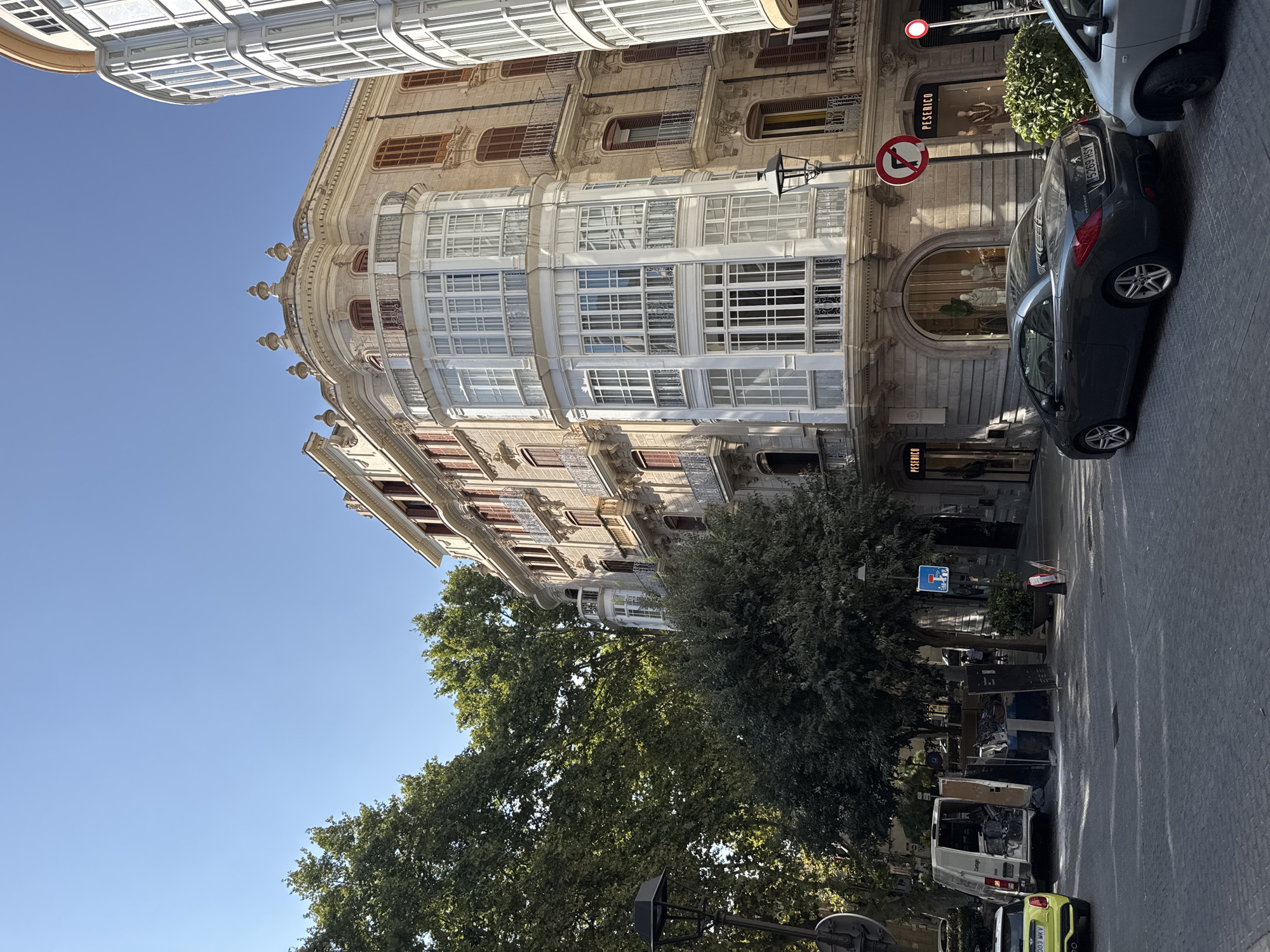 In Palma, Spain, on a clear day, a street scene unfolds in front of a multi-story building with ornate architecture. The building features a prominent bay window with white framing and numerous panes of glass, a decorative balcony with an iron railing, and shutters on some of its windows. Adjacent to this building is another structure with a curved facade and white-framed windows. In the foreground, a dark grey Peugeot convertible is parked on the cobblestone street. To its right, a portion of a silver car is visible. Further down the street, a white delivery van appears to have been emptied, with its contents, including panels and debris, scattered around it. A yellow Mini Cooper is parked to the left of the van. Trees with lush green foliage line the street, and a lamppost stands tall. Traffic signs, including a "no entry" sign and a "no left turn" sign, are visible. The windows of the building display mannequins dressed in clothing, suggesting a retail establishment. The overall impression is one of urban street life with elements of both historical architecture and everyday activities.