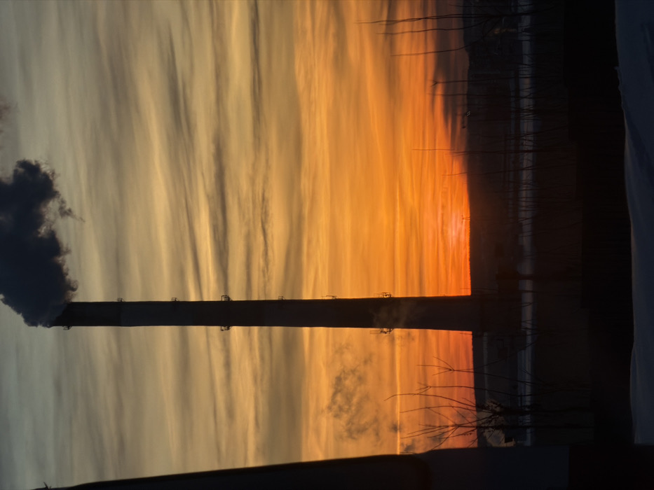 A vertical, long, dark chimney emits smoke against a colorful sky. The sky transitions from orange to light gray with linear cloud formations. Silhouettes of thin trees are visible on the right side. The bottom is obscured by dark objects.