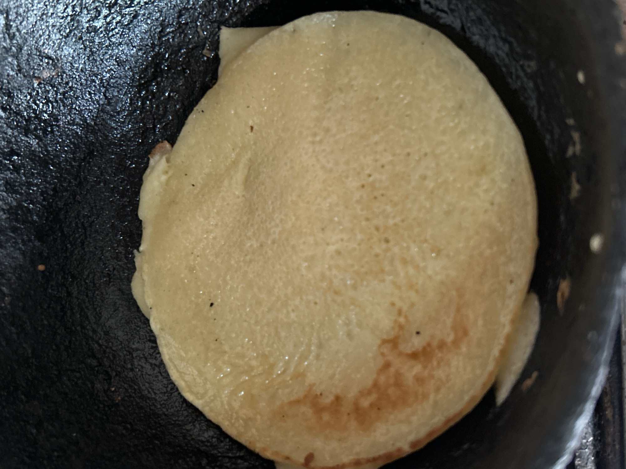 A close-up, overhead shot shows a single, golden-brown pancake being cooked in a dark, non-stick frying pan. The pancake is circular with a slightly uneven edge and appears to be slightly puffed up in the center. The surface of the pancake is textured with small bubbles and has streaks of lighter and darker golden brown, indicating it is cooking evenly. The pan is dark and slightly reflective, with a textured surface. No people or specific location in Santa Ana, Venezuela are visible in the image.