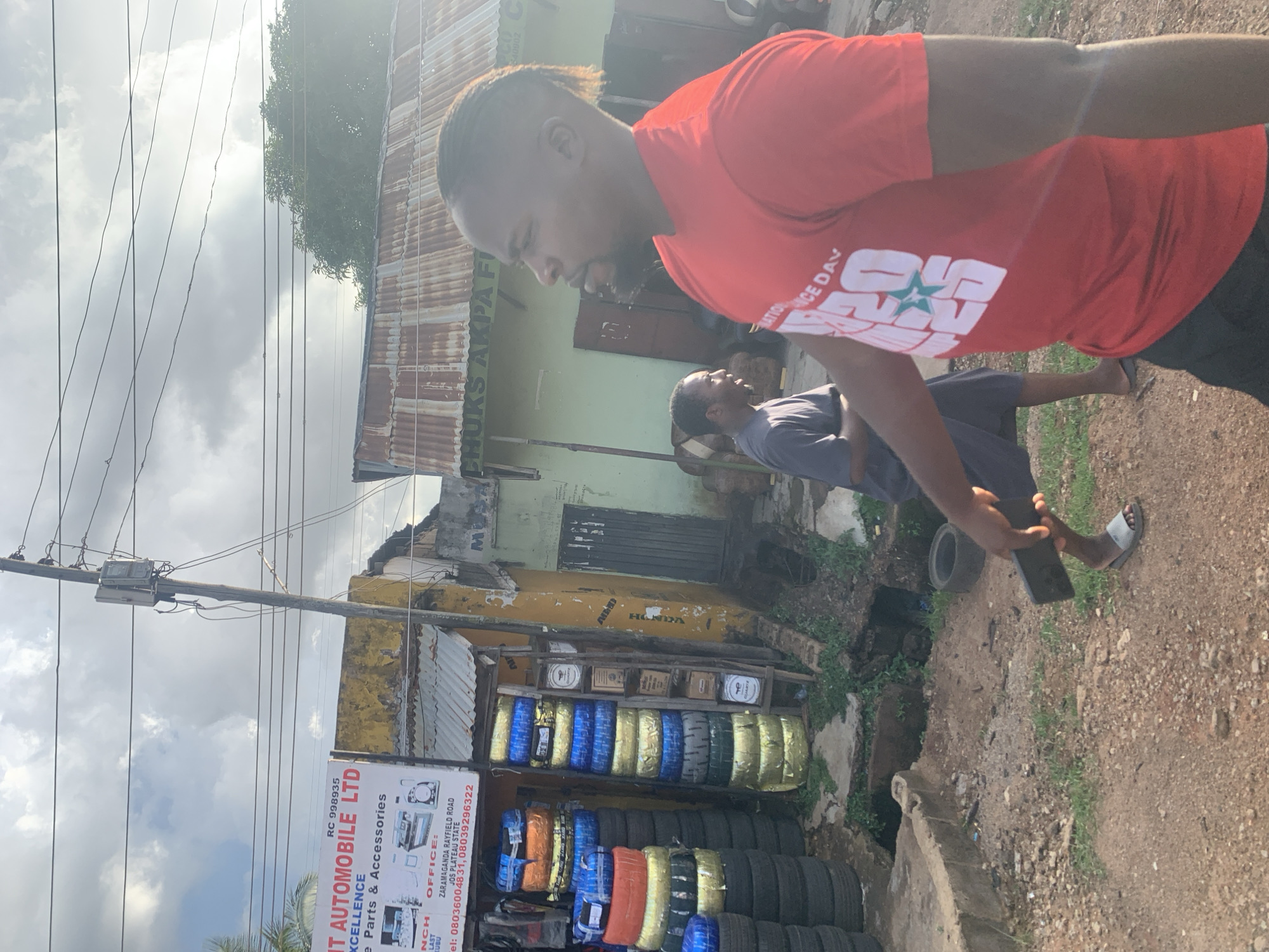 This media file depicts an outdoor scene in Jos, Nigeria, with two men present. One man, wearing a red t-shirt with white lettering and a star emblem, is in the foreground, facing away from the camera and looking downwards. The other man, wearing a dark t-shirt, is further back and also looking downwards. The scene appears to be in front of a shop that sells automobile parts and accessories, as indicated by a sign for "IT AUTOMOBILE LTD" which also lists "Parts & Accessories."
Behind the men, there is a building with a corrugated metal roof and weathered walls. The shop displays stacks of tires, some wrapped in colorful packaging. The ground is rough and dusty, with some overgrown vegetation. A utility pole with electrical wires runs through the scene. The sky is cloudy, suggesting it might be a partly overcast day. The overall impression is that of a busy street in a commercial area. Visible text on signs includes the company name, contact information, and what appears to be a slogan or event related to "NATION ICE DAV." The activity seems to be casual, with the men engaged in looking at something on the ground or a device.