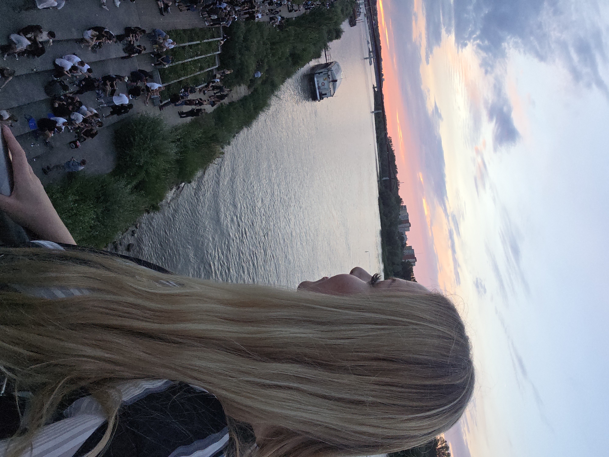 The image shows a young blonde woman with long hair in the foreground, looking to the right at a wide river during sunset. The sky is painted with hues of pink, orange, and blue. On the far bank of the river, a city skyline with buildings and a bridge is visible. Along the near bank of the river, there is a grassy area and a concrete embankment with many people gathered, sitting on steps and the ground. A boat is moored on the river. The scene appears to be a popular public space, possibly a park or waterfront area, in Warsaw, Poland.