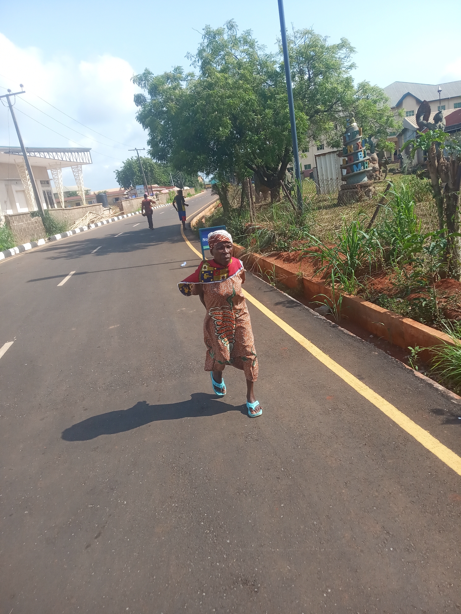 This image captures a bright, sunny street scene in Nsugbe, Nigeria. The main subject is an older woman with dark skin, positioned in the foreground and walking towards the viewer on an asphalt road. She wears a colorful, patterned traditional dress in shades of brown, red, and yellow, a matching headscarf, and light blue flip-flops. She holds a long, slender object, possibly a stick or pole, extended forward with both hands. A prominent shadow of her figure is cast on the road to her left, indicating direct sunlight, likely midday or early afternoon.
Further down the road, two men are also walking away from the camera. The road itself features a solid yellow line on the right side and dashed white lines in the center. On the left side, there is a modern-looking building with a white exterior and an open-frame metal roof structure, alongside a low stone wall and stacks of wooden planks.
To the right of the road, a concrete curb separates the asphalt from an area of red soil and varied green vegetation, including tall grasses. Beyond this, numerous trees with dense foliage are visible, along with parts of other buildings. A distinctive, colorful spiral sculpture, appearing to be made of concrete or stone with stacked letters, stands prominently in the green area on the right. Visible letters on the sculpture include "L," "N," "B," "M," and "H." Another abstract sculpture is partially seen nearby. A blue sign, partially obscured, is visible near the woman with the text "No" legible. The sky is bright with some scattered white clouds.