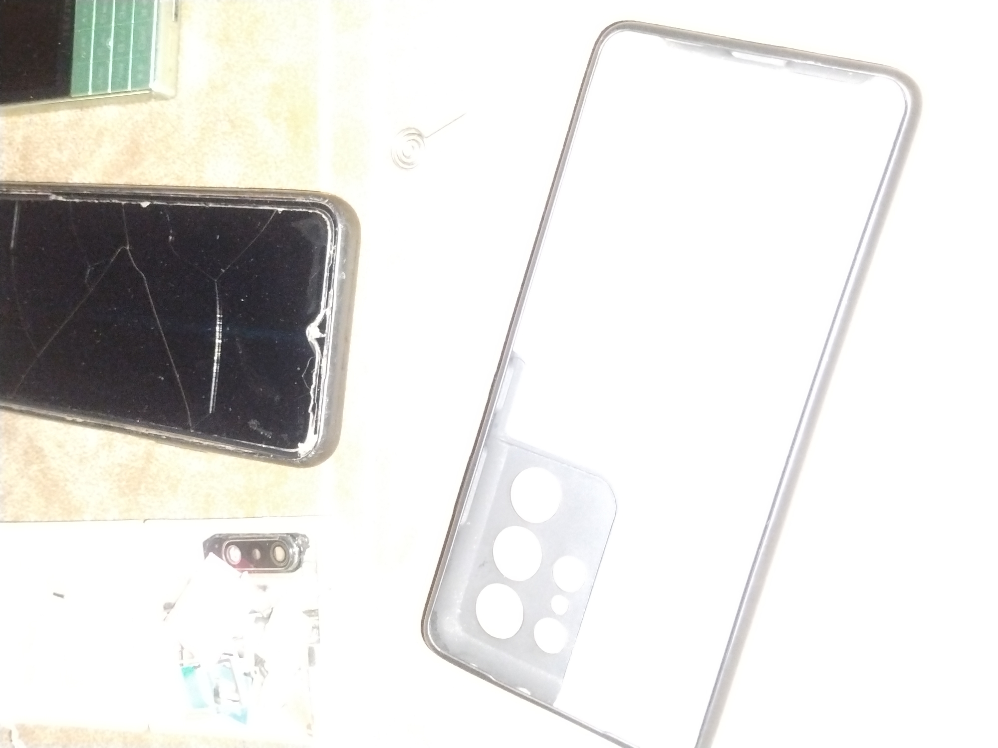 The image shows three mobile phones on a light-colored surface. One phone has a cracked screen. Another phone has a clear case with a camera cut-out design. The third phone, partially visible, appears to be a simple phone. The location, Jos, Nigeria, is not evident in the image.