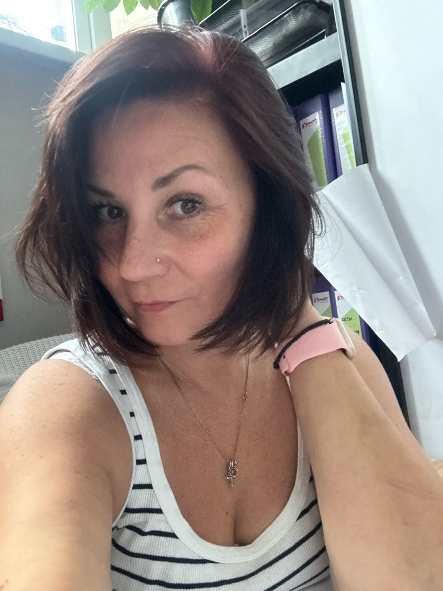 A medium shot captures a woman with shoulder-length dark hair, styled with a slight side part and subtle pink highlights. She is wearing a white tank top with black stripes, a delicate gold necklace with a cross pendant, and a pink smartwatch band. A nose ring is visible on her right nostril. Her left arm is bent, with her hand resting on the back of her neck, drawing attention to the smartwatch. The background is softly blurred, showing a window with natural light, office supplies, and what appears to be binders. The scene suggests an indoor setting, possibly an office or a home office, in Zelenograd, Russia.