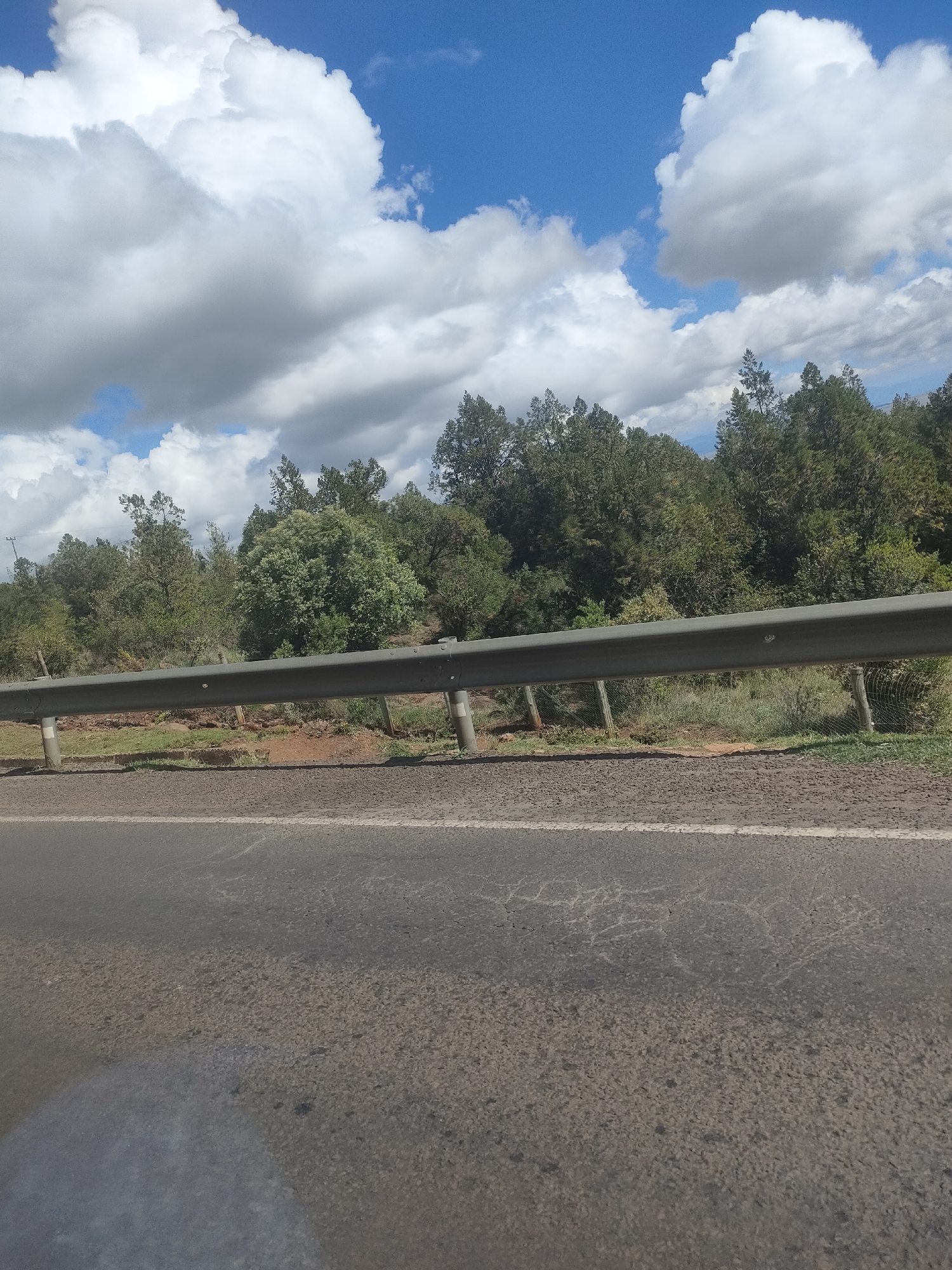 The image depicts a roadside scene in the Kisima ward of Kenya. A two-lane asphalt road with a white line divider is in the foreground. A grey metal guardrail runs parallel to the road, separating it from an area of red soil and green vegetation. Beyond the vegetation, a line of green trees is visible. The sky is blue with scattered white clouds. No people are visible.