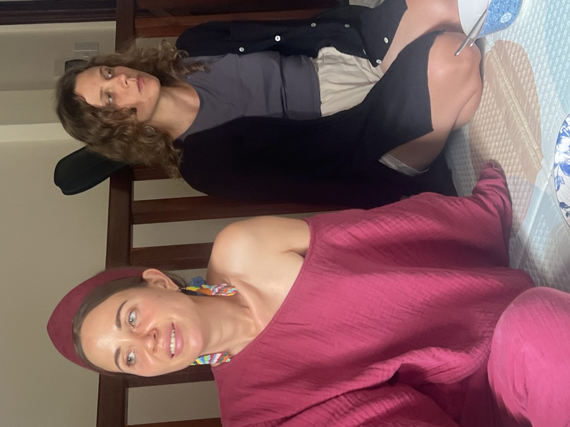 Two individuals are seated on a bed in a room. The person on the left, facing forward, wears a magenta head covering and a matching off-the-shoulder top, adorned with colorful, beaded earrings. The person on the right, looking to their right, has curly brown hair and wears a dark cardigan over a grey t-shirt and light-colored shorts. Both individuals appear to be in a relaxed posture. The background consists of a wooden headboard and a wall. In the foreground, a patterned bedspread is visible, along with parts of blue and white patterned bowls and a utensil. The scene suggests a domestic setting, possibly in Đà Nẵng, Vietnam, given the context.