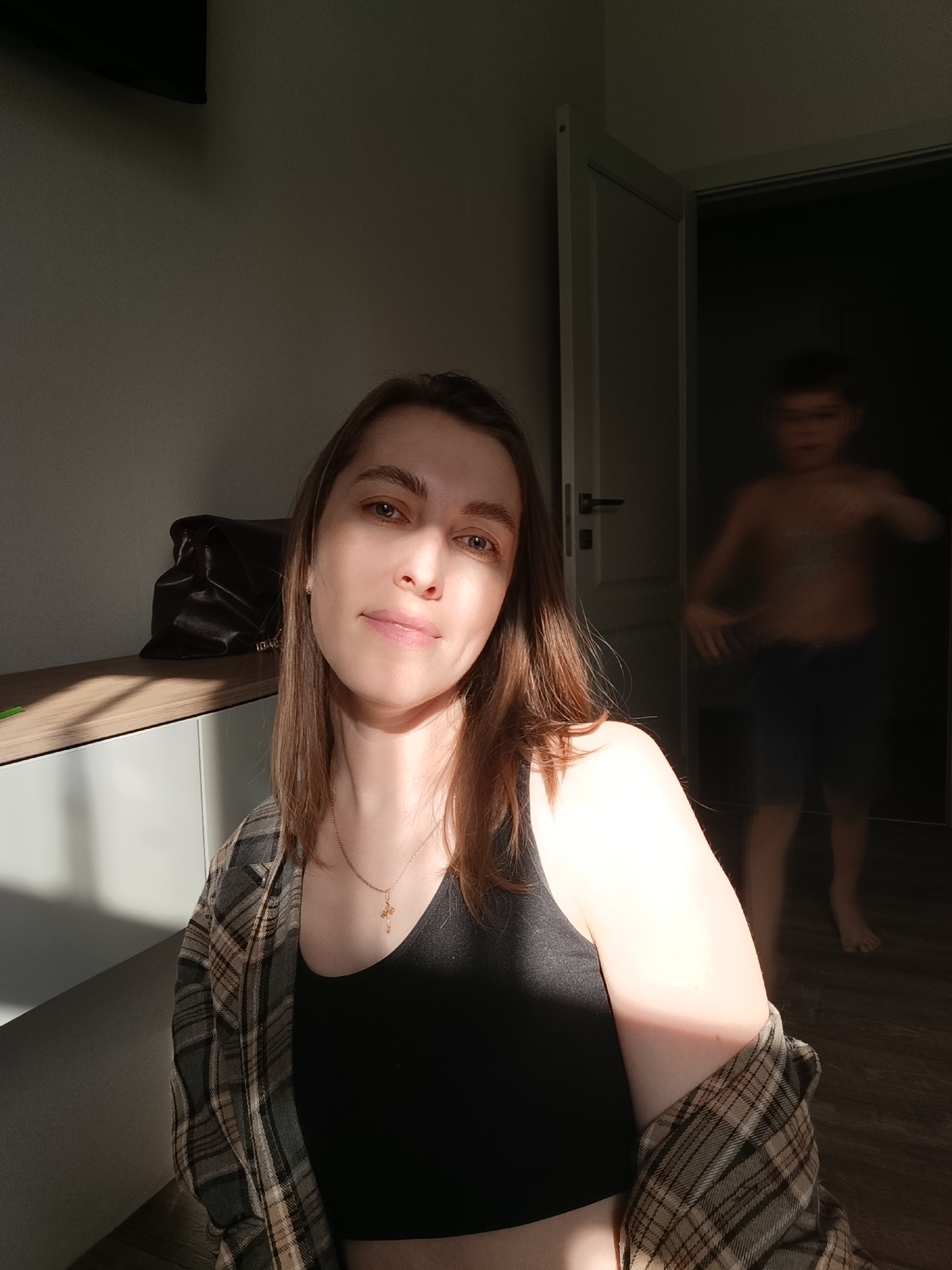A woman is in the foreground, wearing a black tank top and a plaid shirt. She has brown hair and is looking at the camera. In the background, a blurred figure of a child is visible in the doorway. A cabinet with a dark-colored bag on top is to the left of the woman. The scene is illuminated by sunlight. The location is an interior space, possibly a home, in the city of Chaikovsky, Russia.