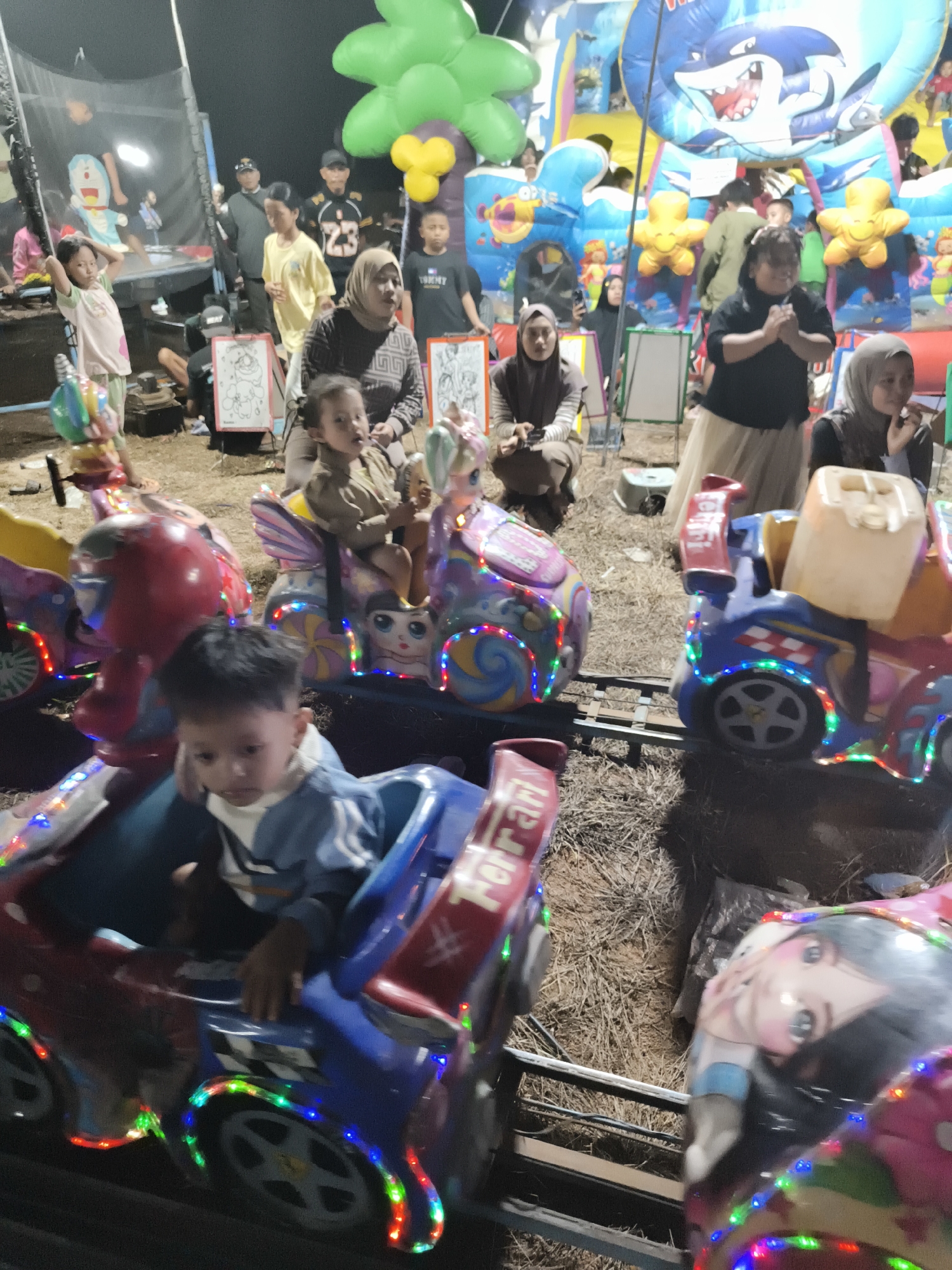 The image depicts an outdoor night scene at what appears to be an amusement area in Selimantan Jaya, Indonesia, likely a local fair or carnival. In the foreground, several children are seated on a miniature train ride. The train cars are brightly colored, decorated with cartoon characters, and adorned with string lights. One young boy in a blue jacket is prominently featured in the closest train car, looking down. Behind him, other children are visible in adjacent cars, some looking towards the camera.
In the midground and background, more people are gathered around various attractions. An inflatable bouncy castle with a shark graphic is visible, as are other inflatable structures that might be part of the amusement setup. Several adults, some wearing headscarves, are present, supervising children or observing the activities. The ground is covered with straw or dry grass, typical of such outdoor events. The overall atmosphere is lively and festive, characterized by the presence of amusement rides, crowds, and nighttime lighting.