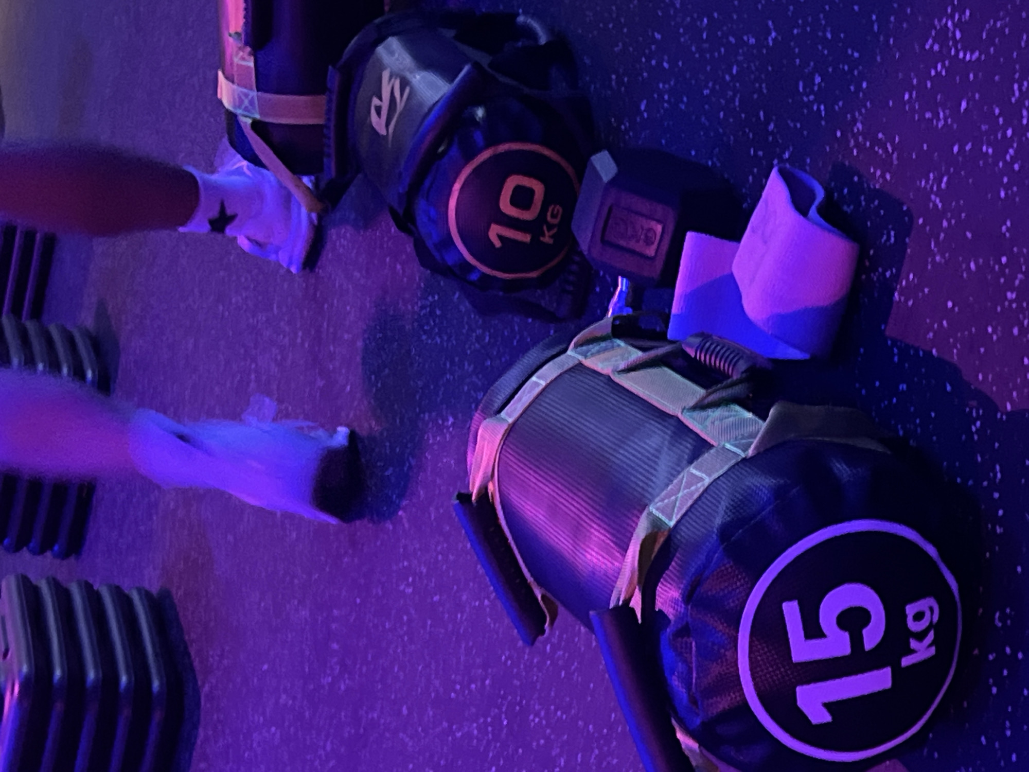The image captures a ground-level view of fitness equipment and partial figures in a gym or fitness studio, illuminated by strong purple and blue ambient lighting. Two individuals are present, evidenced by a person's forearm and hand (wearing a white glove or sleeve with a black star design) near a 10kg weight bag, and another person's legs and feet (wearing light-colored socks/shoes) positioned near stacked weight plates.
The central subjects are two weight bags, likely sandbags or similar functional training tools. One bag is black with a circular yellow outline, featuring the text "10 KG" prominently in orange. Above it, a partially visible logo or text appears to start with "SK". Another, larger black weight bag is seen in the foreground on the right, displaying "15 KG" in white lettering within a circular white outline. Both bags have visible straps. A black dumbbell is positioned between the two weight bags. To the far left, several dark, rectangular weight plates are stacked. A pink or light-colored accessory, possibly a wrist strap or cushion, rests near the dumbbell.
The floor is dark with a speckled pattern, typical of gym flooring. The vibrant purple and blue lighting creates a dynamic, atmospheric setting, suggesting an energetic workout environment or a specific type of fitness class, possibly in the evening or night. No specific activity is clearly depicted beyond the presence of people and equipment in a workout space.