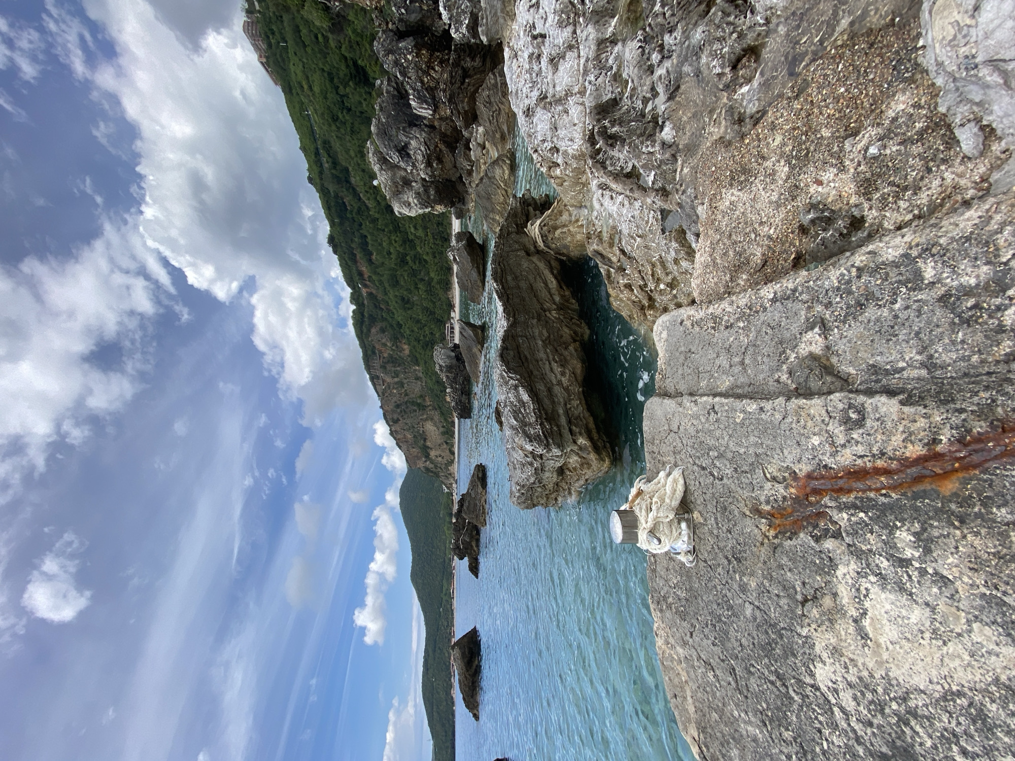 The image shows a view of the Adriatic Sea from a rocky shoreline, likely in or near Prijevor, Montenegro. The coastline is rugged with rock formations extending into the water. A cliff face covered in green vegetation is visible in the background. In the foreground, a concrete or stone platform is visible with a mooring cleat and a coiled rope attached to it. The sea is calm with clear blue water. The sky is partly cloudy with patches of blue.
