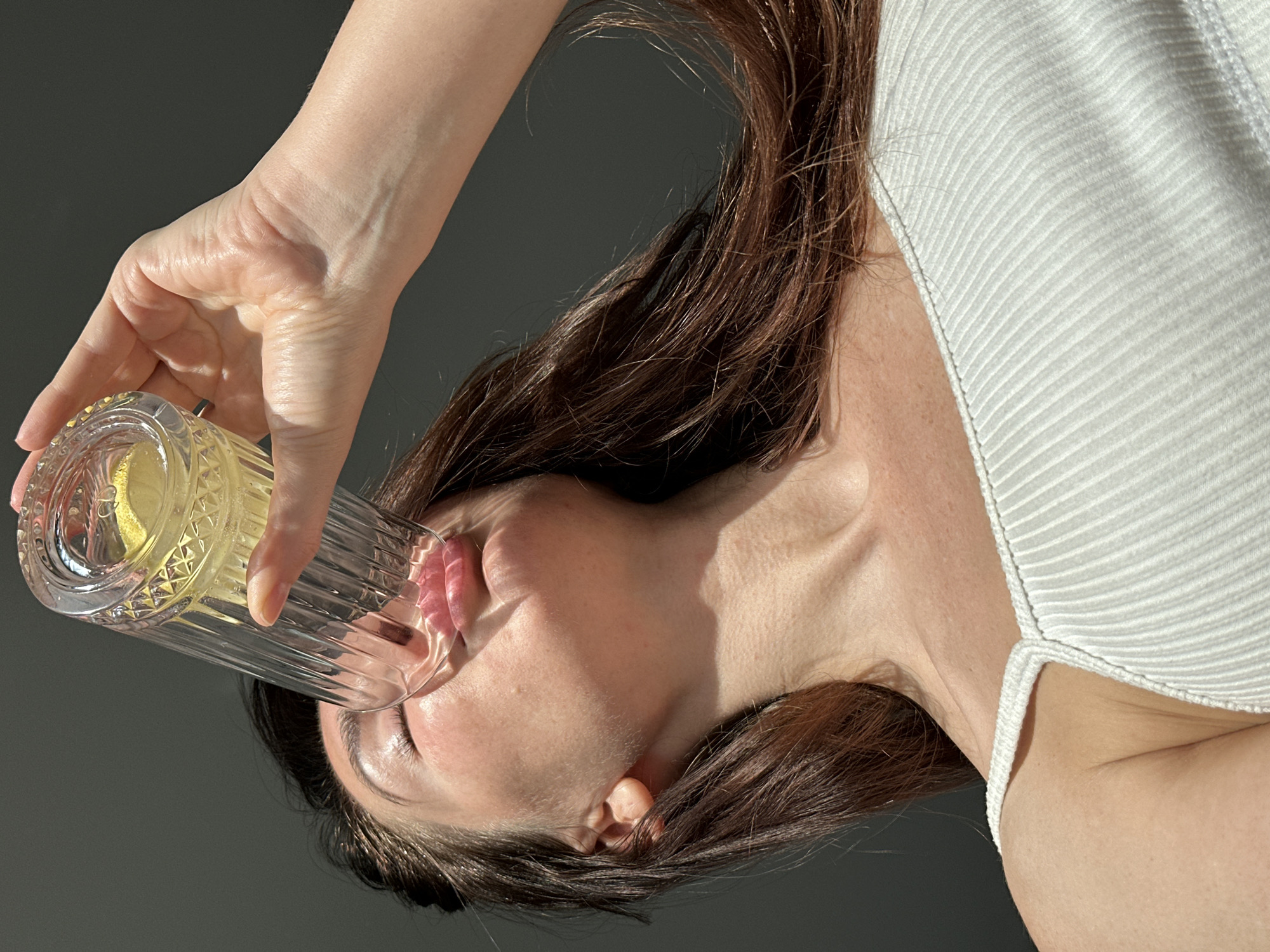 A woman is drinking from a faceted glass, presumably containing lemonade or another yellow beverage. She is wearing a white ribbed tank top. Her hair is brown. The background appears to be a solid gray color. There is no verifiable evidence to connect this image to the city of Chaikovsky, Russia.