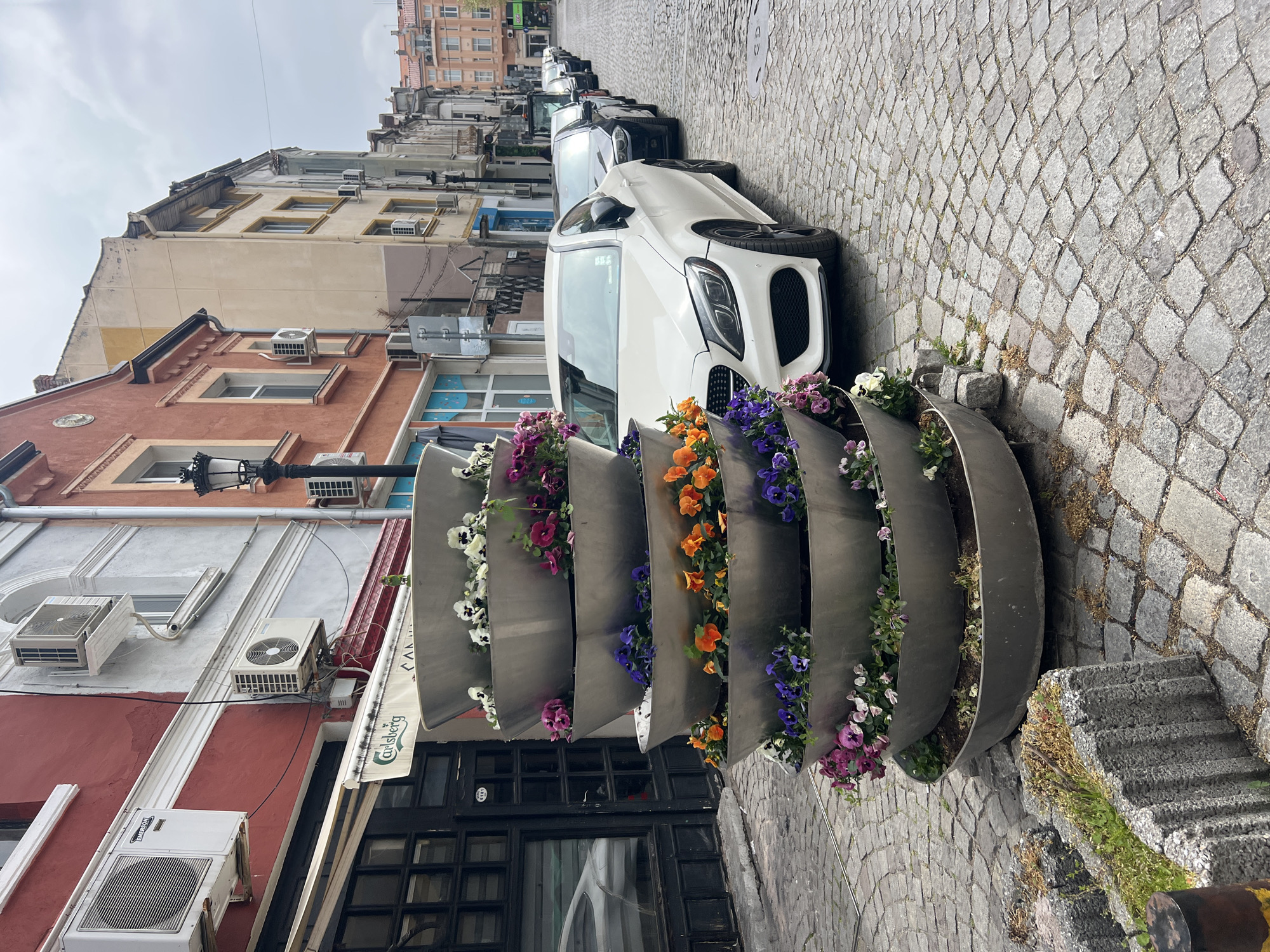 The image depicts a street scene in Plovdiv, Bulgaria. Cobblestone paving covers the street. A series of curved, metal planters filled with various colored pansies line the edge of the street. A white car is parked alongside the planters. Several other cars are visible parked further down the street. Buildings of varying heights and colors line the other side of the street, many featuring air conditioning units mounted on their facades. The sky is overcast. A Carlsberg awning is visible above a building's entrance.