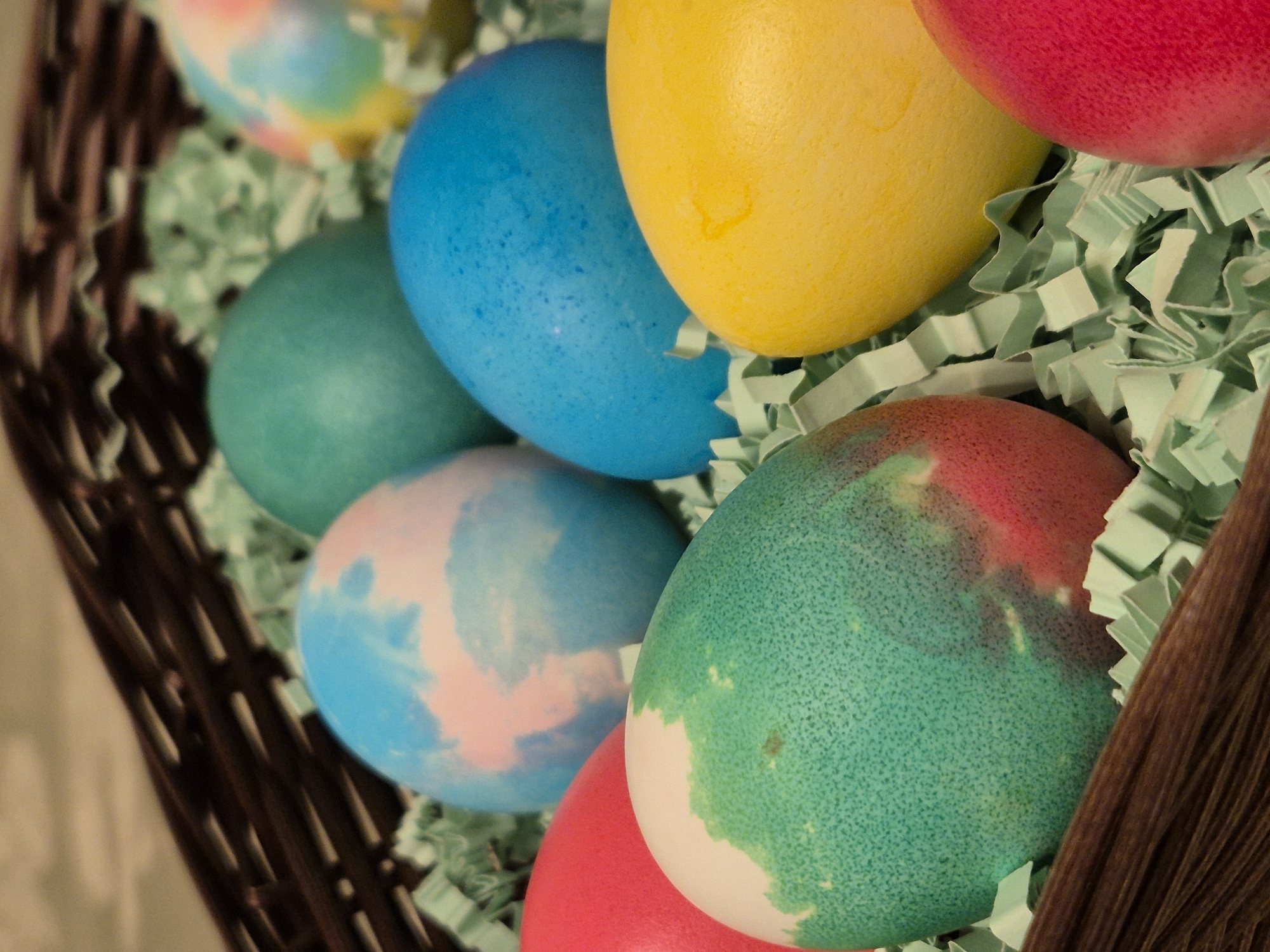 A close-up shot displays a collection of dyed Easter eggs nestled within a woven basket. The eggs feature various colors including red, yellow, blue, and green, with some displaying marbled or multi-toned effects. They are arranged among light green paper Easter grass inside the basket. The focus is on the eggs and their arrangement, with the basket's weave visible in the periphery. No information suggests any connection with the city of Varna, Bulgaria. There are no people depicted in the frame, and the scene focuses primarily on the decorated eggs and their presentation.