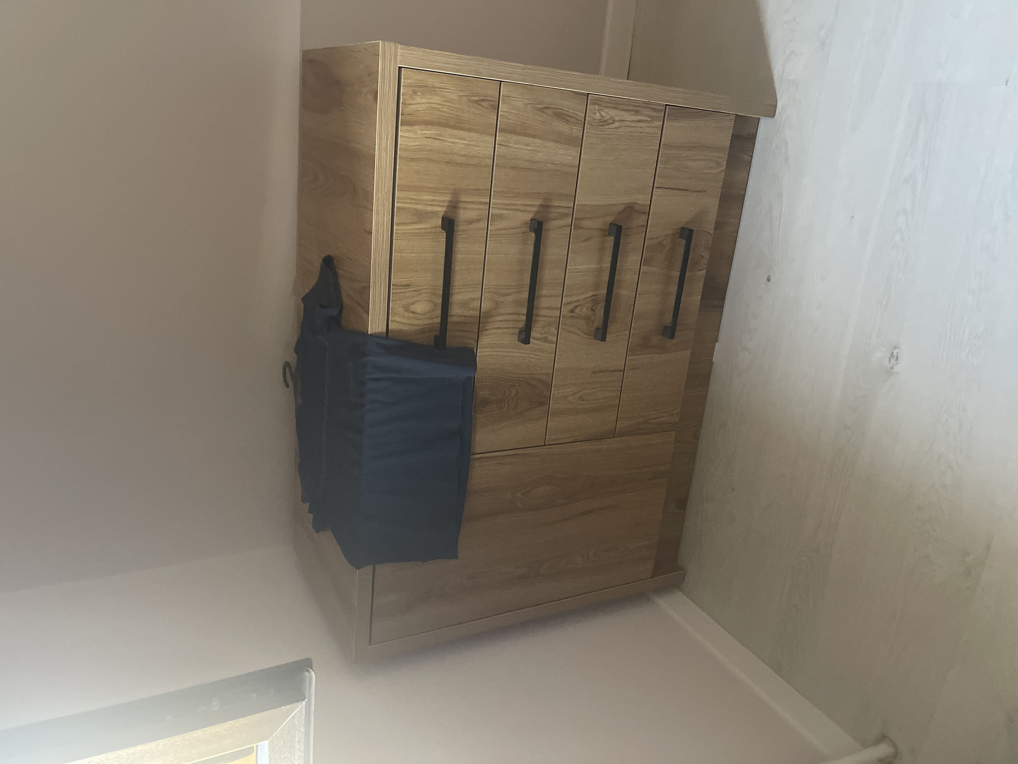 A dark blue t-shirt is draped over the top of a light brown wooden dresser. The dresser has four drawers, each with a black horizontal handle. The dresser is positioned in a room with light pink walls and a light grey wooden floor. There is a window on the left side of the image, partially obscured by light. This scene is located in the city of Qarshi, Uzbekistan.