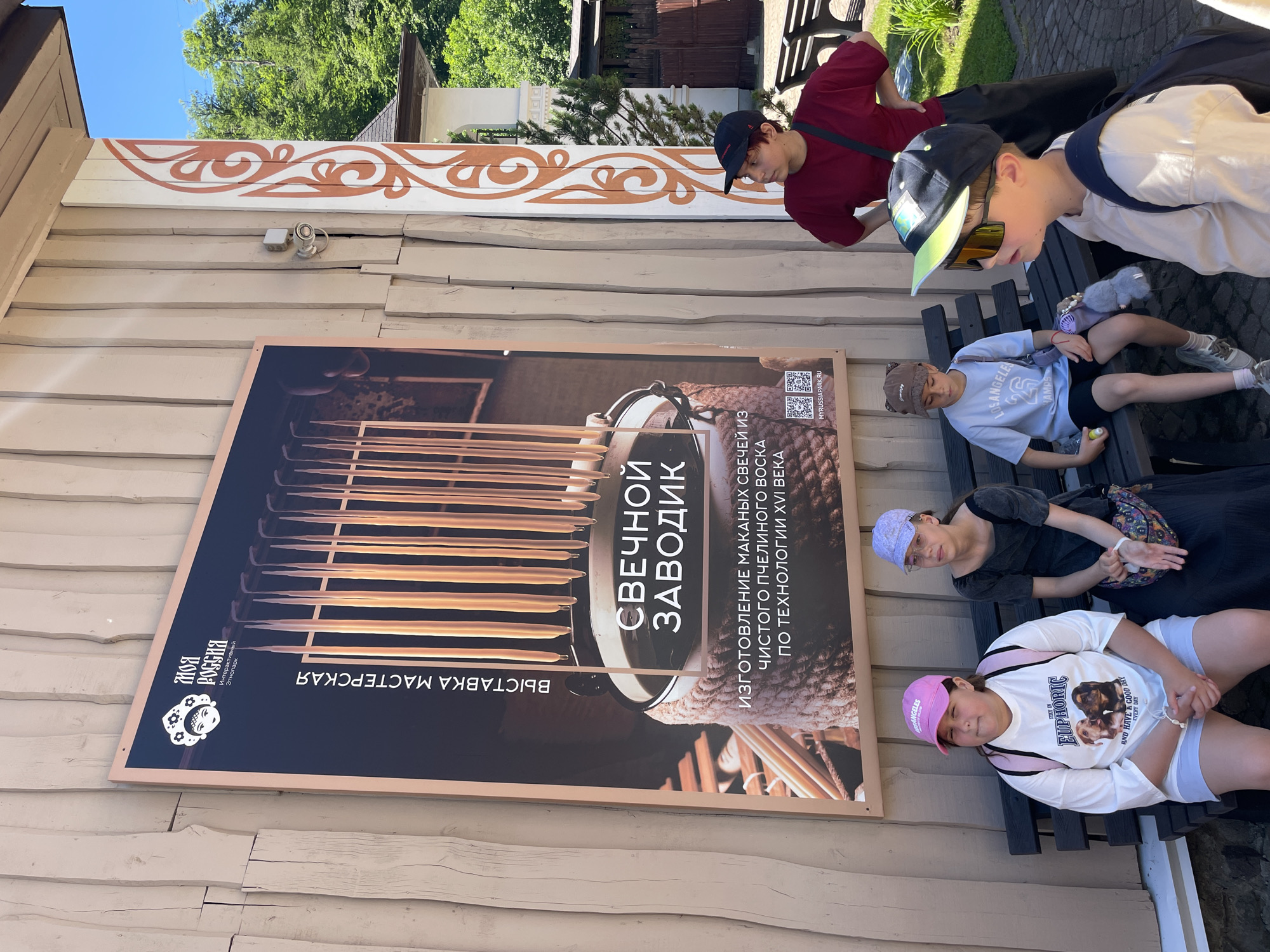 In Estosadok, Russia, an outdoor scene features a large poster on a wooden building. The poster advertises "My Russia Interactive Park" and depicts a candle-making workshop. In front of the poster, a group of four children and one adult are present. Two young girls and two young boys are seated on a wooden bench. An adult woman stands behind the bench, looking to the right. The children are dressed in casual attire, including t-shirts, shorts, and hats. One girl wears a pink baseball cap, while another wears a purple cap. The boys are also wearing baseball caps, one with a bear-themed hat and the other with a lime green brim. One boy is wearing sunglasses. The environment appears to be sunny with greenery visible in the background.