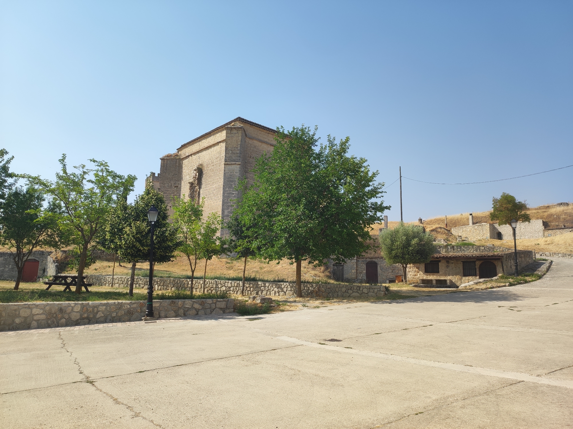 This image captures a view of the Church of San Miguel in Ampudia, Spain. The church, a substantial stone building with a pitched roof, is partially visible on the left side of the frame, showing its textured stone facade and a large, arched window opening. In front of the church, a variety of trees with lush green foliage are present, with a picnic table situated in the grassy area to the left. A black lamppost stands prominently in the foreground, casting a shadow on the concrete ground. To the right of the church, a small, rustic stone building with a tiled roof and dark wooden doors and windows is visible, nestled into the side of a gently sloping hill. The landscape is characterized by dry, golden grass on the hillside, with stone retaining walls and structures dotting the terrain. The sky above is a clear, bright blue, indicating a sunny day. The scene depicts a tranquil, historical setting within the town.
