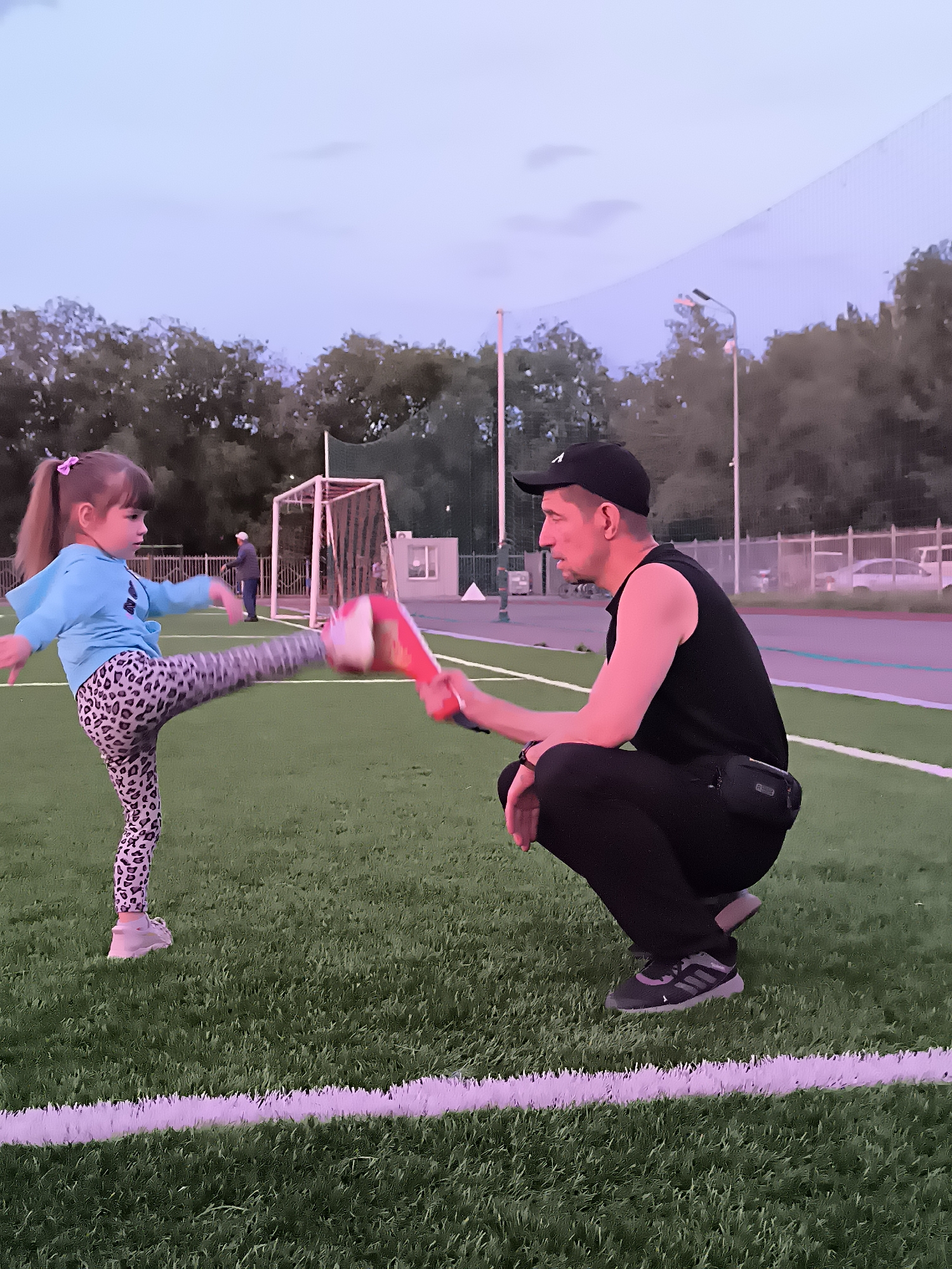 In this image, a young girl with a pink hair tie in her ponytail is practicing kicking a padded target held by a man. The girl is wearing a blue hoodie and patterned leggings, while the man is dressed in a black sleeveless shirt, black pants, and a black baseball cap. They are on a green artificial turf field, likely a sports field, with white lines marking boundaries. In the background, there's a soccer goal, trees, and a fence. The sky is a pale twilight blue, suggesting it is early morning or late evening. The lighting appears to be artificial, possibly from stadium lights, casting a pinkish hue on the scene. The overall atmosphere seems to be one of training or recreation, with a focus on the interaction between the man and the child. The location context provided is Qarağandı city administration, Kazakhstan. There is a white letter 'A' visible on the man's baseball cap.