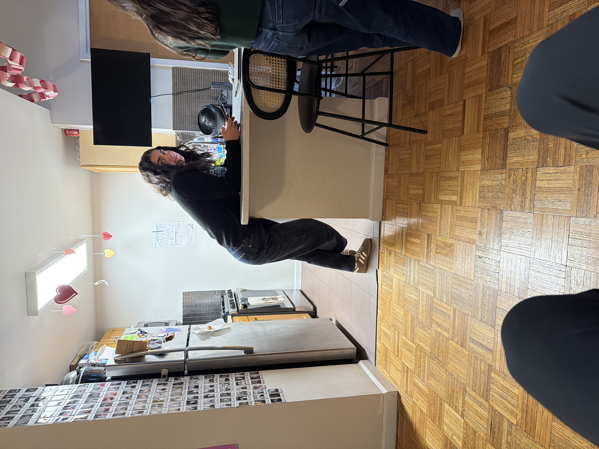 The image shows an indoor scene, likely a kitchen, in what appears to be an apartment. The floor is wooden parquet. A woman with dark hair stands leaning against a white kitchen island, with a black top. Behind her is a stove. On the wall are pictures of people. Other items visible in the kitchen include a refrigerator covered in photos, a toaster, a microwave, and a ceiling light fixture with heart-shaped decorations hanging from it. Another person is sitting on a chair at the island. The location is not definitively identified as New York City. No specific forensic details are visible.