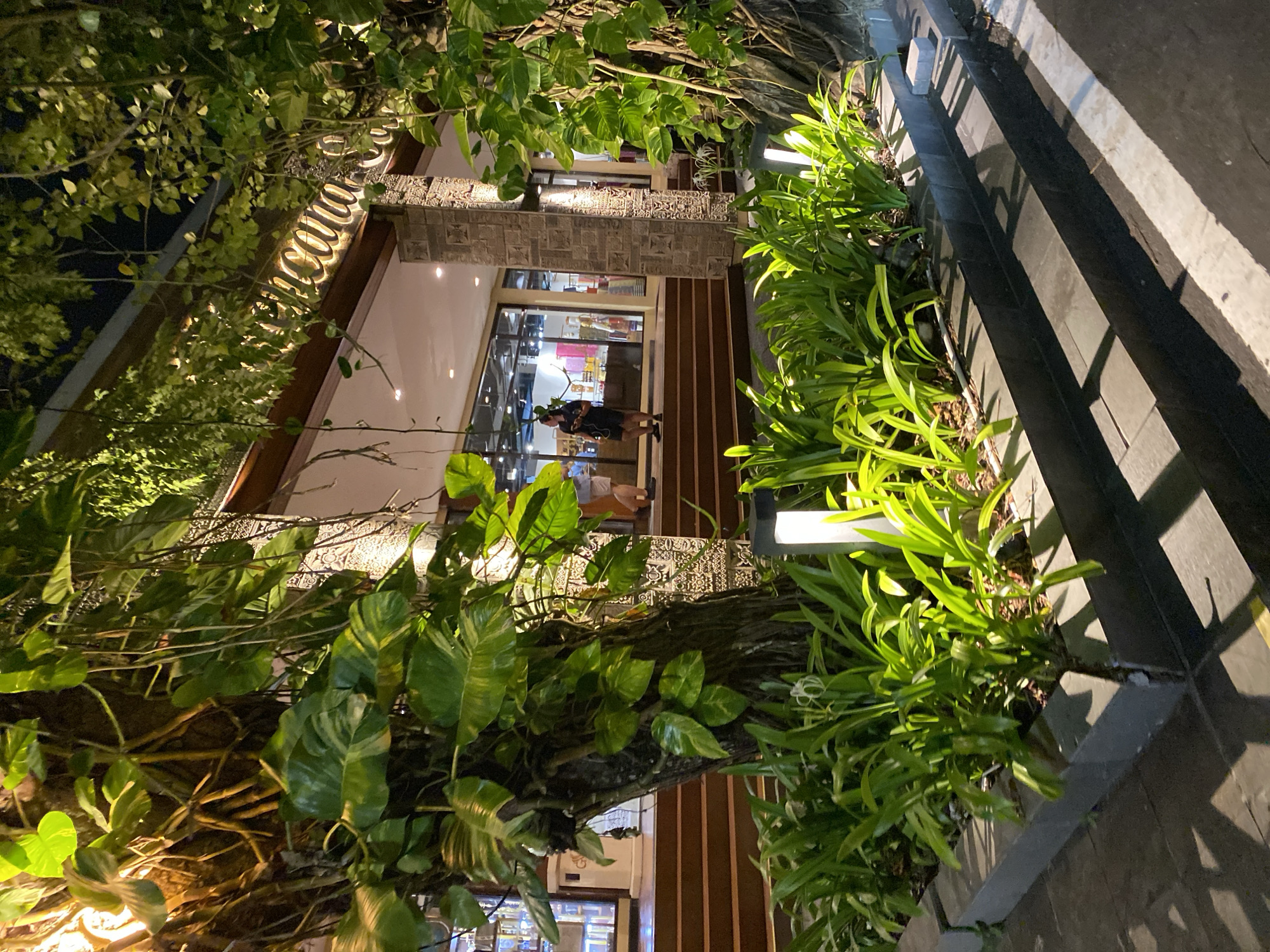 The image shows an exterior view of a building at night in Kuta Selatan, Indonesia. The building has a sign with illuminated text that reads "Kencana" in the upper right portion of the image. The entrance to the building is set back and features large glass windows. A wide set of wooden stairs leads up to the entrance. Lush green plants, including large-leafed vines climbing trees and a planter box filled with smaller leafy plants, are prominent in the foreground and frame the scene. Several modern, rectangular outdoor lights are placed amongst the smaller plants, casting a warm glow. A tree trunk with vines wrapped around it is positioned in the left foreground. In the mid-ground, two individuals are visible. One person, wearing dark clothing, stands on the stairs facing left. Another person, partially obscured, is also on the stairs. The ground in the foreground appears to be paved, with a white line marking a section of the path. The overall atmosphere is tropical and inviting, with the warm lighting accentuating the greenery.