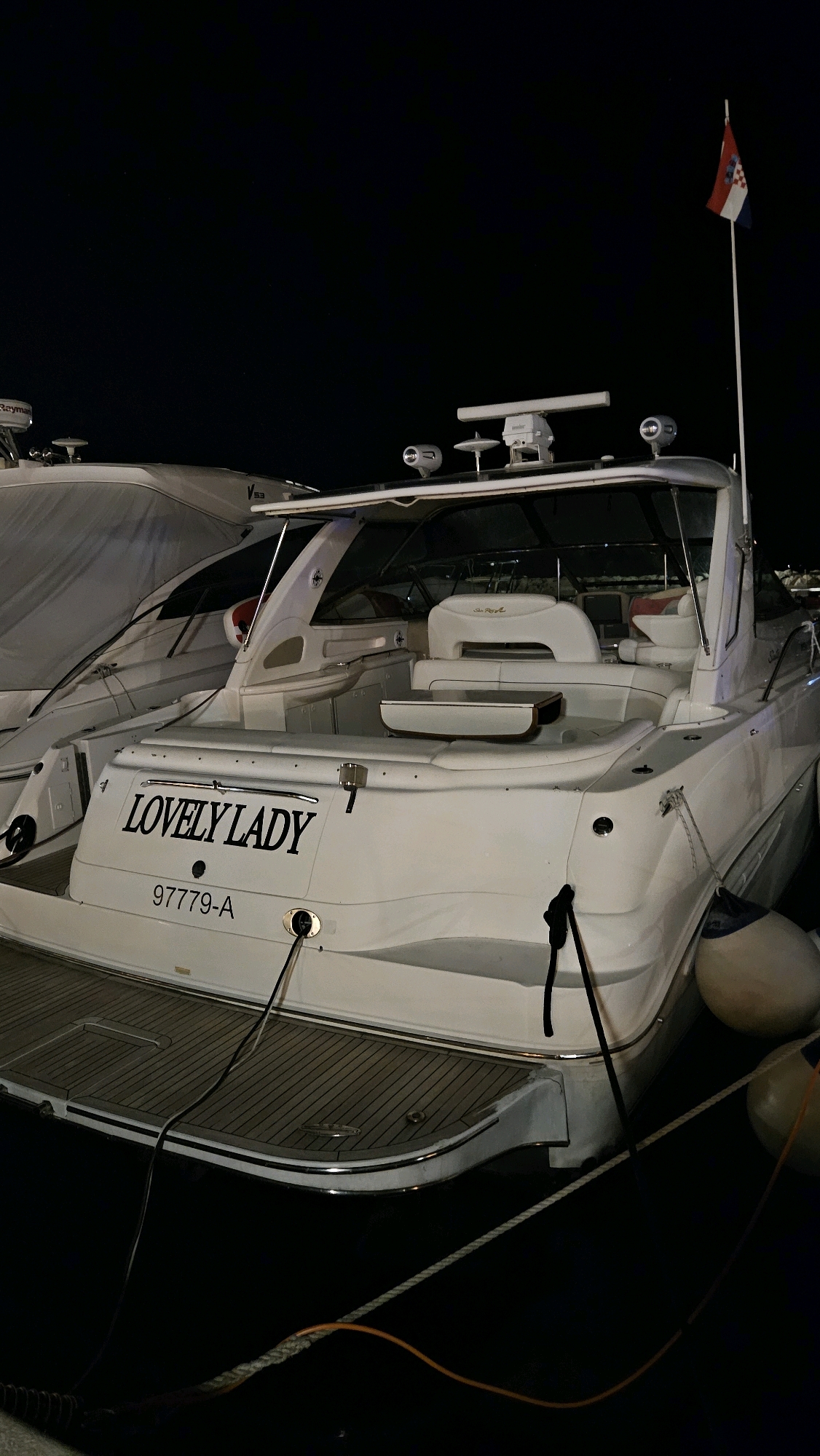 A white motor yacht named "LOVELY LADY" with registration "97779-A" is moored at night in the harbor of Općina Tučepi, Croatia. The yacht is white with black lettering and a grey trim. It features a radar, navigation lights, and a Croatian flag flying from a white mast at the stern. There are two large white fenders attached to the side of the yacht. The yacht is tied to the dock with ropes, and electrical cables are connected to the vessel. Another yacht, partially covered with a white tarp, is docked alongside to the left. The scene is dimly lit, suggesting it is nighttime, and the water is calm.