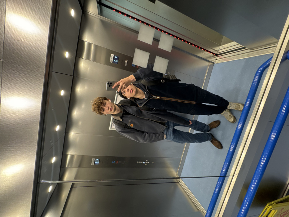 The image depicts two males in a mirror selfie taken inside an elevator, located in Newcastle upon Tyne, United Kingdom. The elevator interior features brushed stainless steel walls and a light blue floor. The male on the left, with curly brown hair and white earbuds, wears a grey work-style jacket, blue jeans, and brown shoes, with a dark backpack visible. The male on the right, wearing a dark beanie, a dark zippered jacket over a dark hoodie, dark trousers, and light-colored boots, holds a silver smartphone with both hands, angled upwards to capture their reflection. He also has white earbuds and a dark patterned backpack. Visible on the elevator walls are control panels: one on the left displaying a floor indicator "S" with an upward arrow, and another on the right featuring square buttons and a vertical red LED strip. A blue handrail is present in the lower foreground, below which a partial yellow "WET FLOOR" sign is reflected.