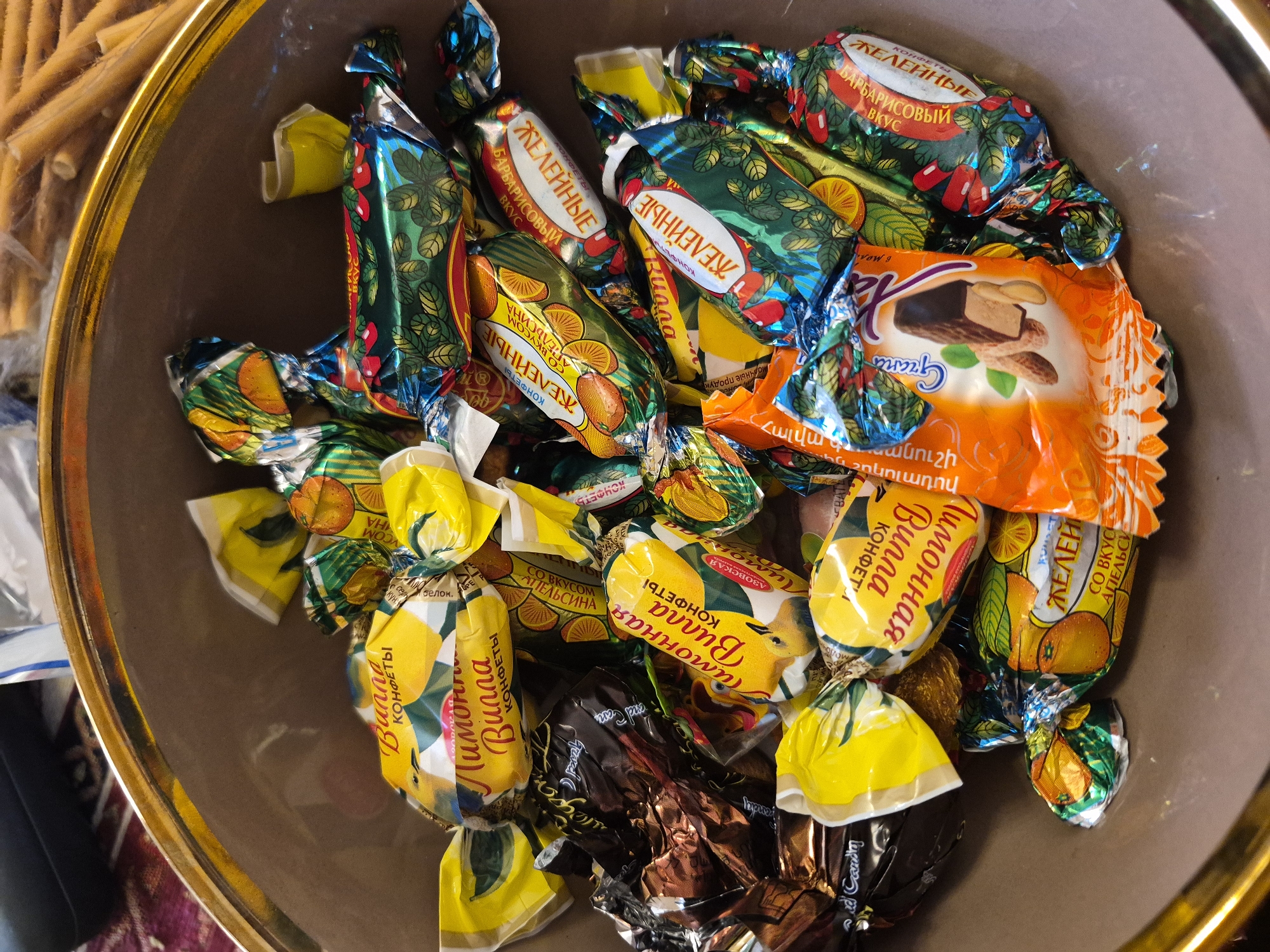 A brown bowl with a golden rim is filled with an assortment of individually wrapped candies, viewed from a high angle. In the blurred background, to the upper left, long, light-colored snack sticks or waffle rolls are visible.
The candies feature diverse wrappers:
- Many are in shiny green and blue foil, often decorated with leaves, berries, or orange slices. Russian text "ЖЕЛЕЙНЫЕ КОНФЕТЫ" (Jelly Candies) is prominent. Some specify "СО ВКУСОМ АПЕЛЬСИНА" (with orange flavor), while others indicate "БАРБАРИСОВЫЙ ВКУС" (barberry flavor).
- Numerous candies are wrapped in bright yellow foil, typically adorned with lemon imagery. The Russian text "КОНФЕТЫ Лимонная Вилла" (Candies Lemon Villa) is clearly visible, suggesting a lemon-flavored variety. The word "АЗОВСКАЯ" (Azov) appears on some of these.
- A prominent orange wrapper depicts a chocolate bar with peanuts. This wrapper features the brand "Granto" (Գրանտո) and the Armenian text "Հալվա շոկոլադով" (Halva with chocolate), indicating a chocolate-covered halva candy.
- A few candies are wrapped in darker, possibly brown or bronze, foil, with some English text like "Candy" partially visible.
The scene suggests an indoor setting, likely on a surface with a patterned textile, as hinted by the background. The multilingual text on the wrappers (Russian, Armenian, English) points to a diverse regional market, consistent with the provided context of Yerevan, Armenia. The image showcases a collection of sweets, likely intended for consumption or sharing.