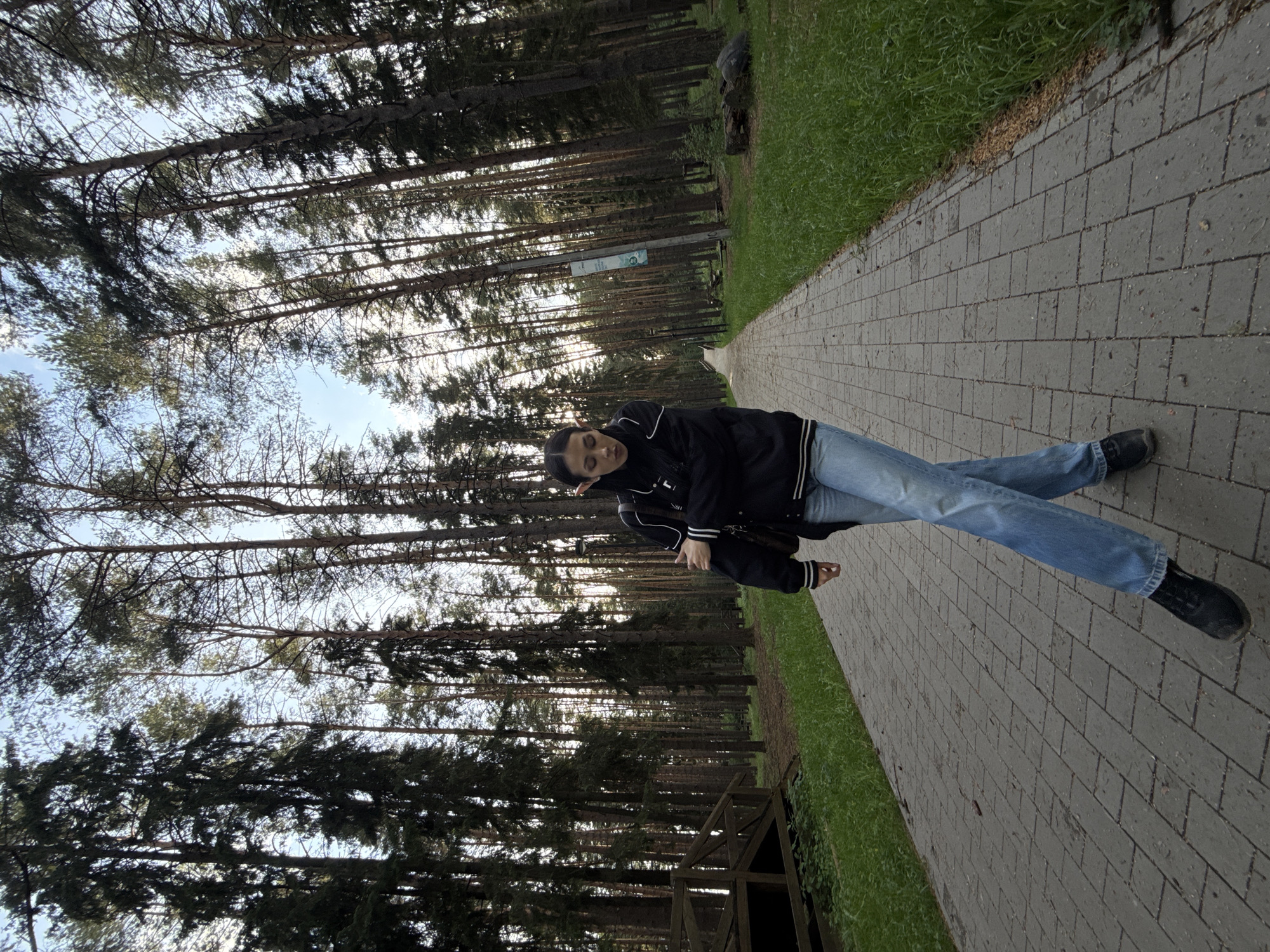 A young woman lies on a gray brick path in a park-like setting. She is wearing a dark jacket with white trim on the sleeves, blue jeans, and dark shoes. The path is bordered by green grass on one side and a dense forest of tall, thin trees on the other. The trees appear to be evergreens. The sky is visible through the trees in the background. The location is likely a park or green space within Votkinsk, Russia.