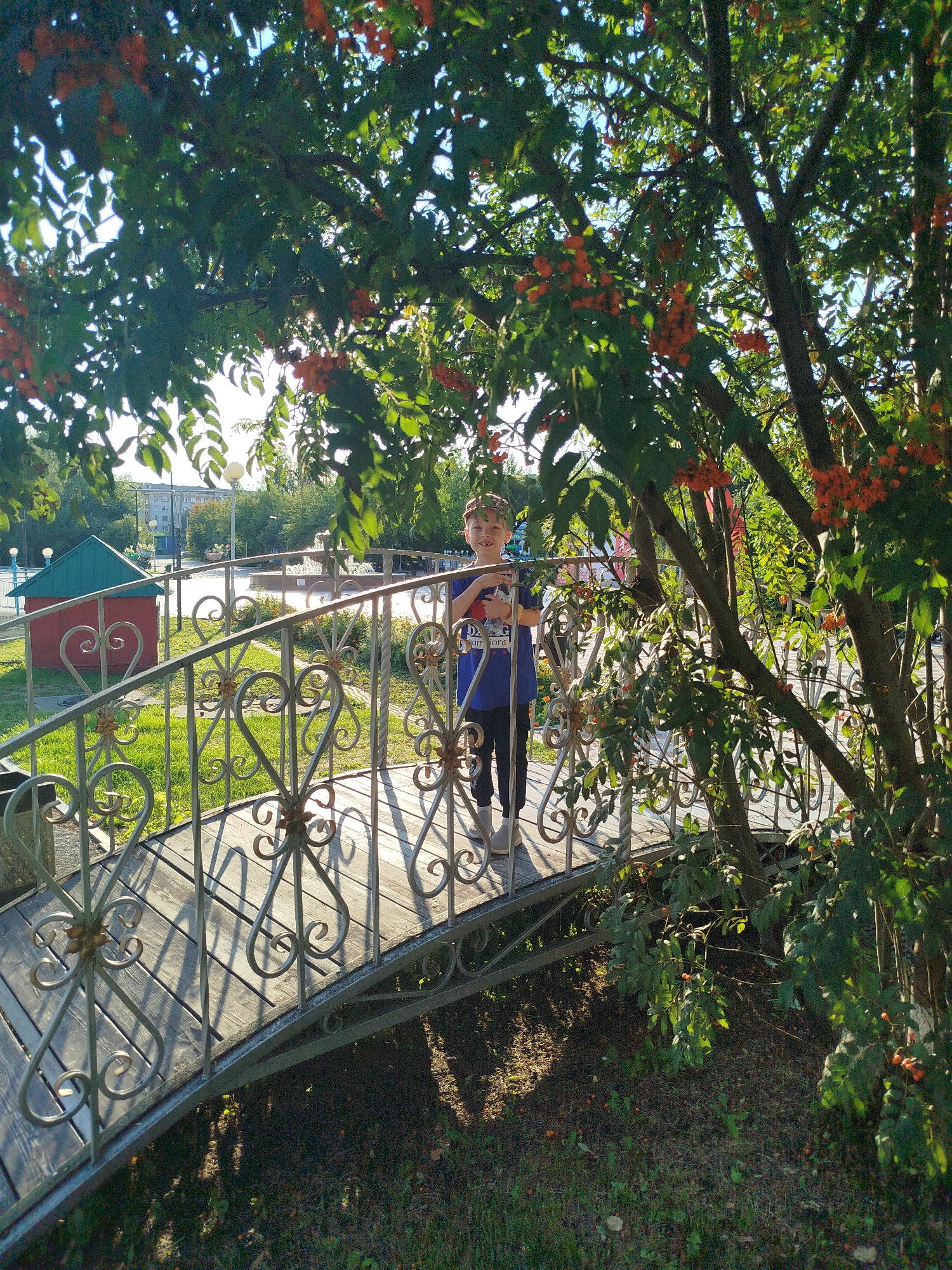 A young boy stands on a decorative metal and wooden bridge in a park in Yalutorovsk, Russia. He is wearing a blue t-shirt and black pants, and he is smiling at the camera. The bridge is surrounded by trees with orange berries. In the background, there is a small red building with a green roof and a fountain. The sun is shining, casting shadows on the bridge and the ground. The scene appears to be set in late summer or early autumn.