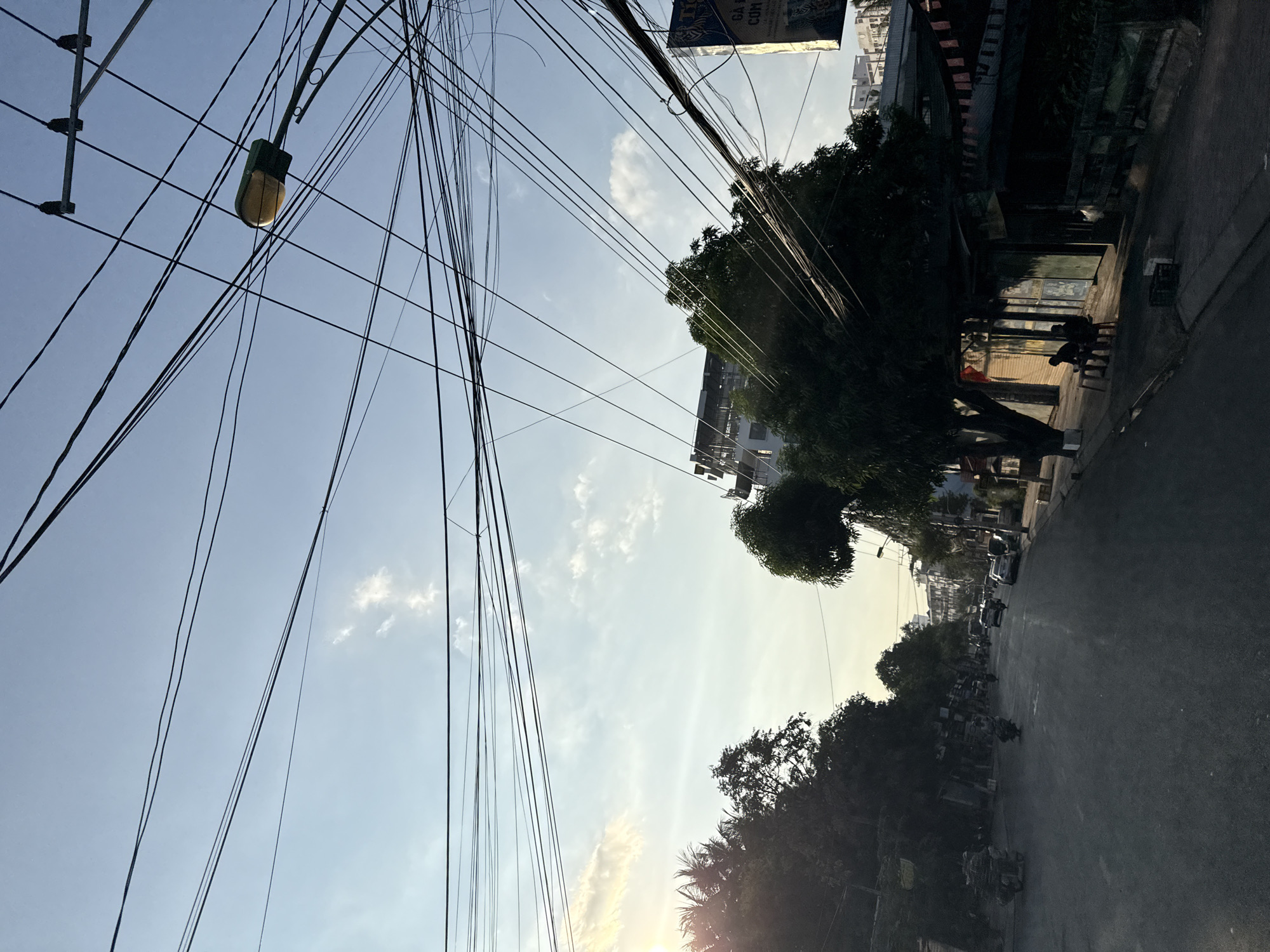 The image depicts a street scene in Bac Nha Trang Ward, Vietnam, likely during the late afternoon or early evening, as indicated by the low sun angle and long shadows. The sky is a pale blue with scattered white clouds. A prominent feature of the image is the dense network of overhead power lines crisscrossing the frame, intersecting with a streetlamp.
The street itself is paved and appears to be a two-way road, with some vehicles, including motorcycles and cars, visible in the distance. The left side of the street is lined with trees and some low-rise structures, while the right side features buildings, some with storefronts. Two individuals are seated on chairs outside a building on the right side of the street, casting long shadows. The overall impression is of a typical urban street in Vietnam, characterized by its dense infrastructure and daily life.