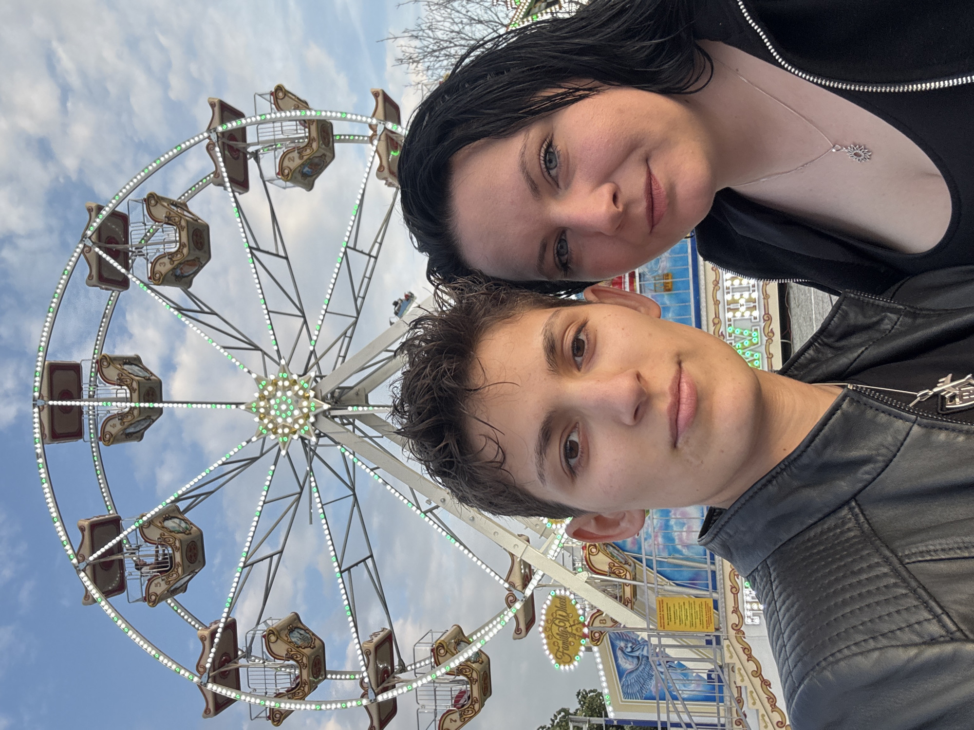 This image is a selfie-style portrait taken in Elin Pelin, Bulgaria, featuring two individuals positioned in the foreground against the illuminated backdrop of a Ferris wheel and a fairground.
The individual on the left has short, dark, textured hair and brown eyes, looking directly at the camera with a neutral expression. They are wearing a black leather jacket with quilted shoulder details over a black top, and a silver necklace with a pendant.
The individual on the right has long, dark hair and blue eyes, also looking directly at the camera with a neutral to slightly smiling expression. They are wearing a black zippered hoodie over a dark top, with a silver necklace featuring a sun-shaped pendant.
In the background, a large Ferris wheel with a light grey metal framework is prominently displayed. It features numerous ornate gondolas, primarily beige and maroon, some of which are occupied by people (e.g., gondola '5' shows occupants). The Ferris wheel is extensively illuminated with small LED lights arranged in alternating green and white patterns along its circumference and forming a starburst design at its center. Below the Ferris wheel, parts of other fairground structures are visible, including an illuminated sign that reads "Family Wheel" and a decorative panel depicting a winged horse (Pegasus) in a blue sky. A metal railing is present in the lower background. The sky above is partially cloudy blue, indicating a late afternoon or early evening setting, consistent with the active illumination of the rides. Bare tree branches are visible on the far right.